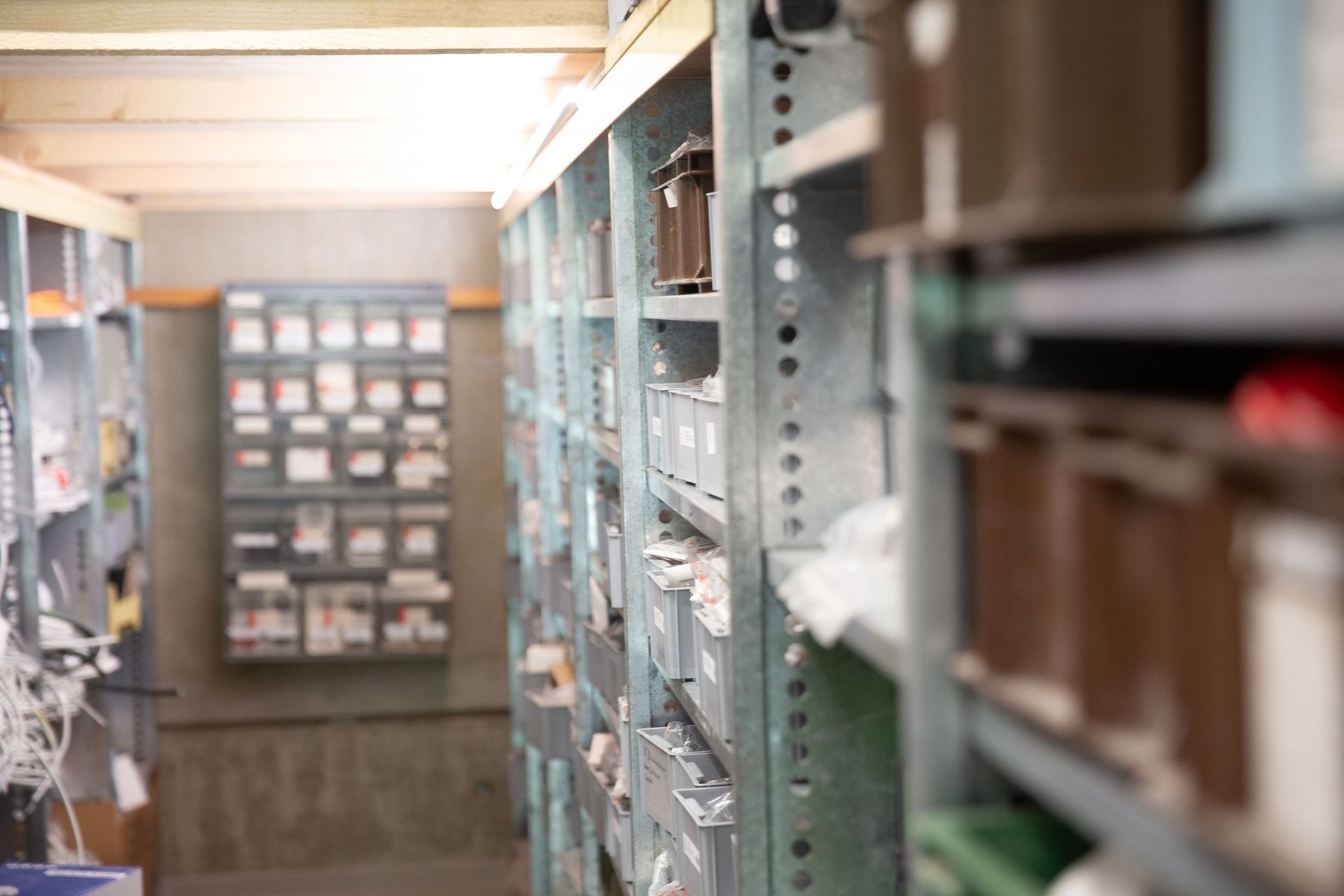 Regale voller Kisten und Vorräte in einem schwach beleuchteten Lagerraum. Der Fokus liegt auf den Regalen und ihrem Inhalt.