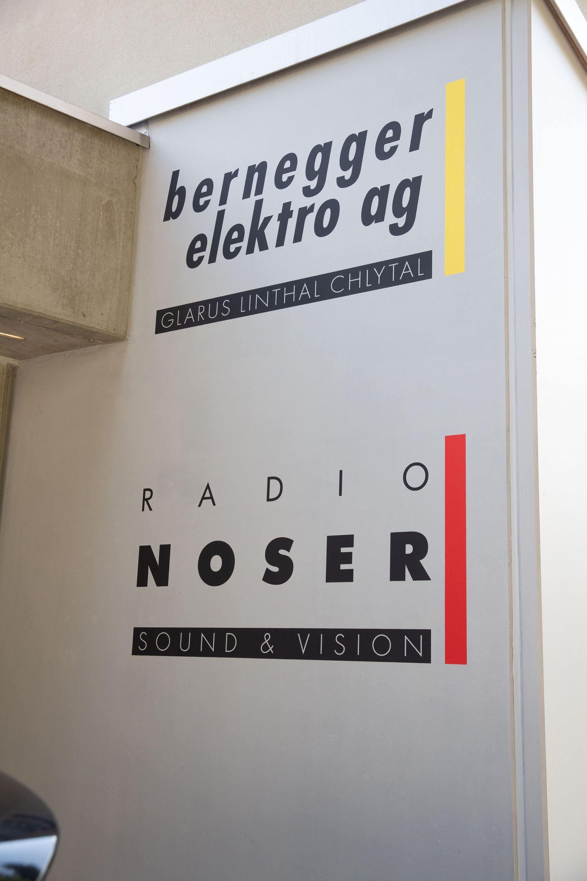 Schild mit den Logos „bernegger elektro ag“ und „radio noser“, grauer Hintergrund mit gelben und roten Streifen.