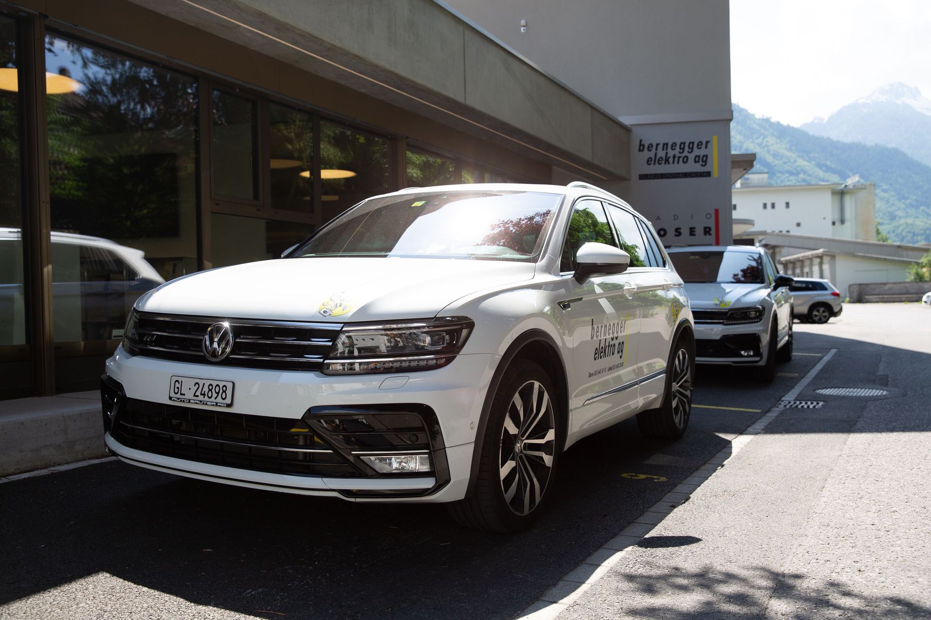 Ein weißer Volkswagen SUV parkt vor einem Gebäude mit Bergen im Hintergrund. Ein weiteres Auto steht dahinter.