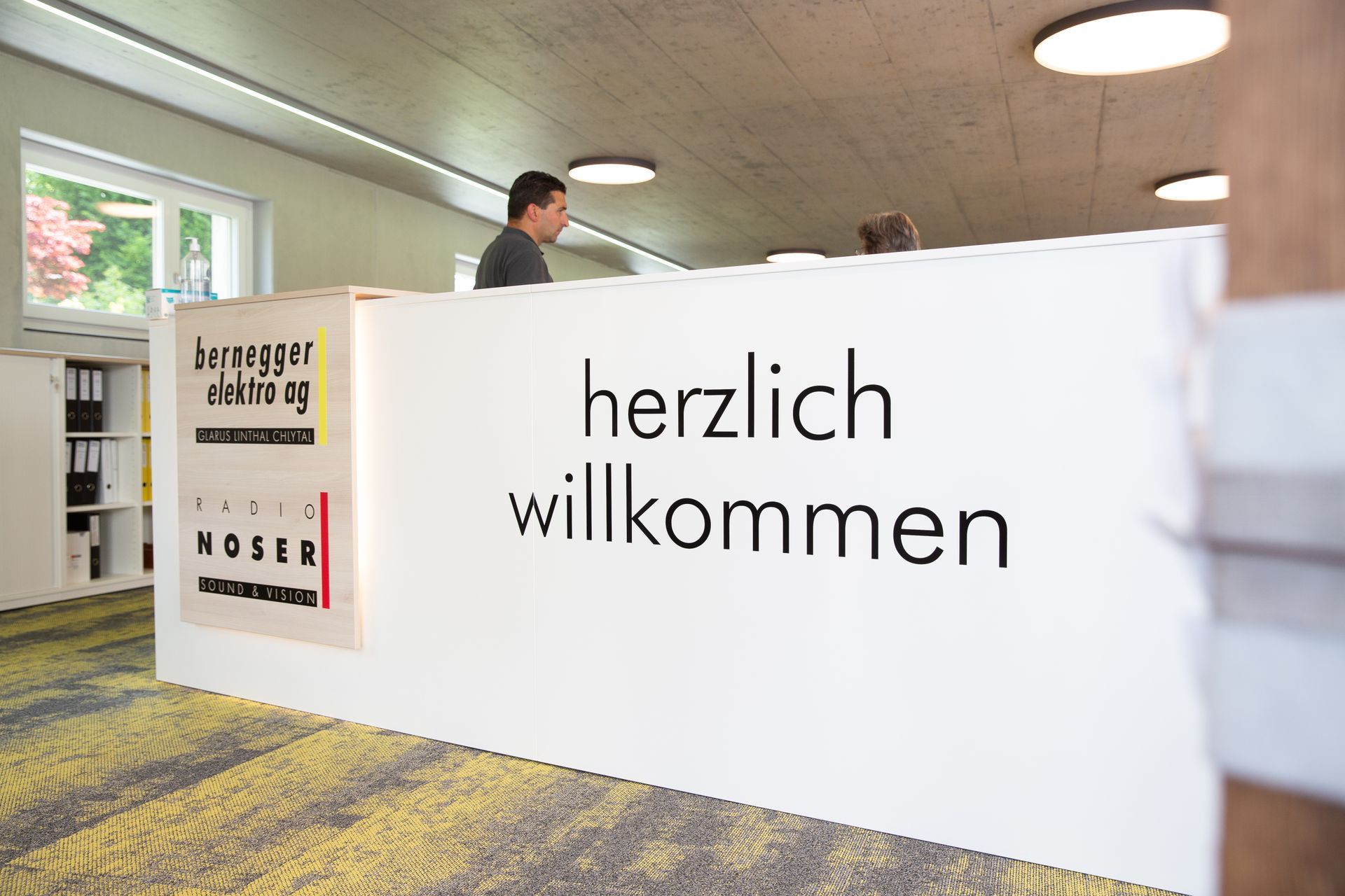 Rezeptionstresen mit der Aufschrift „Herzlich willkommen“. Ein Mann steht hinter dem Tresen.