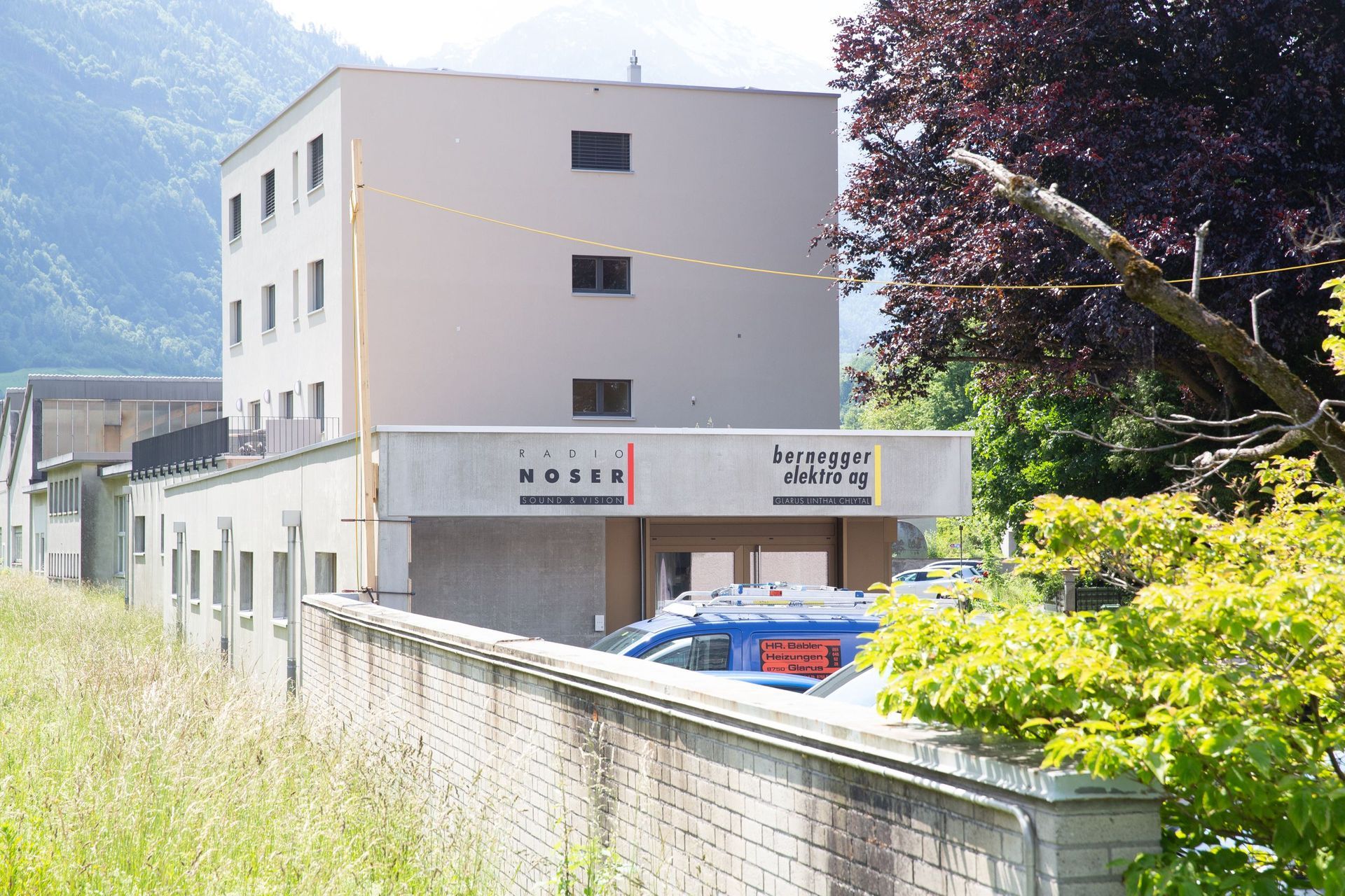 Ein hellbeiges Hotelgebäude mit einem Schild mit der Aufschrift „Hotel Moser“, im Hintergrund ein Berg und davor überwucherte