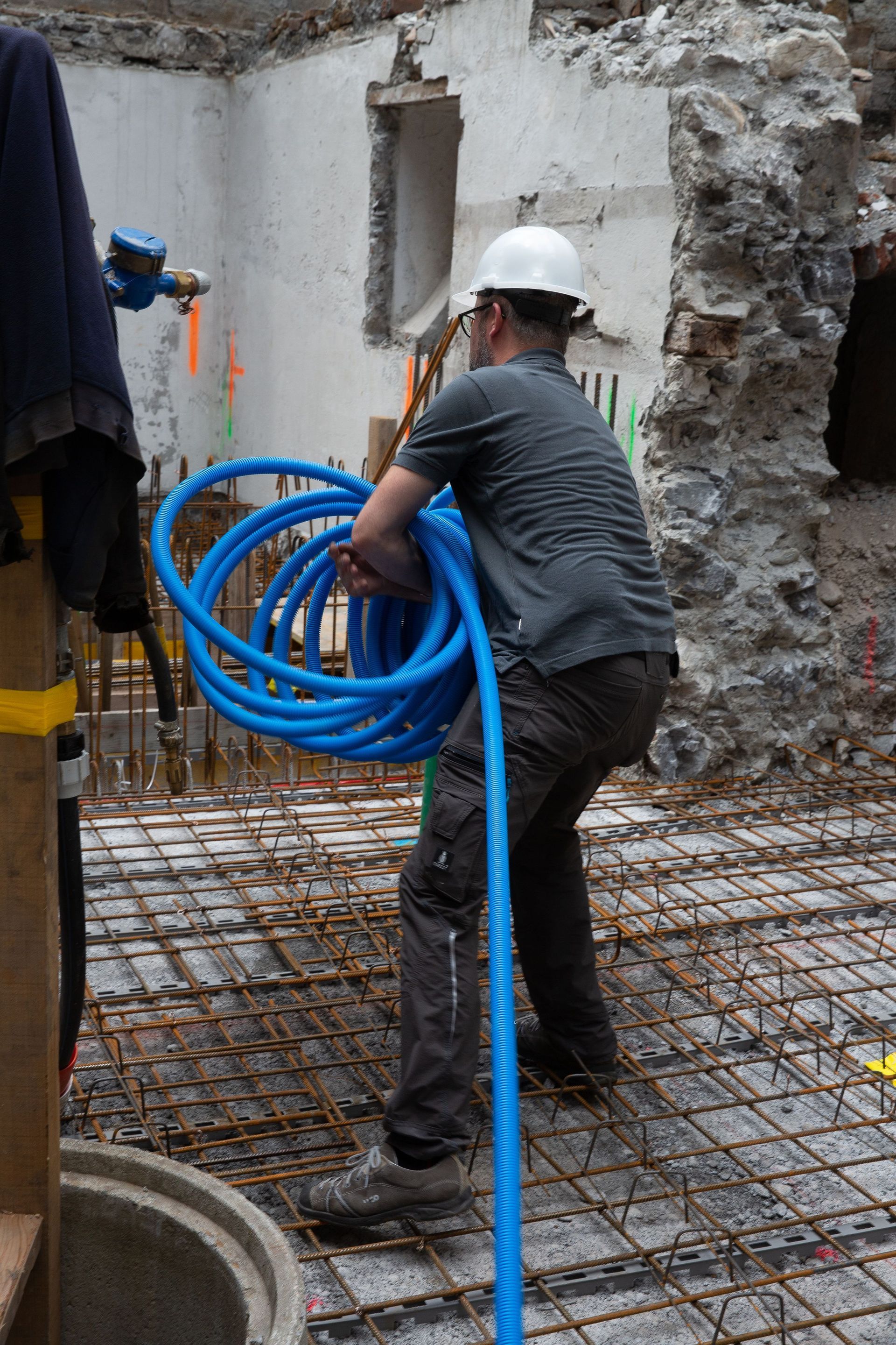 Bauarbeiter trägt eine Rolle blauer Rohre auf einer Baustelle mit Betonwänden und Bewehrungsstahl.