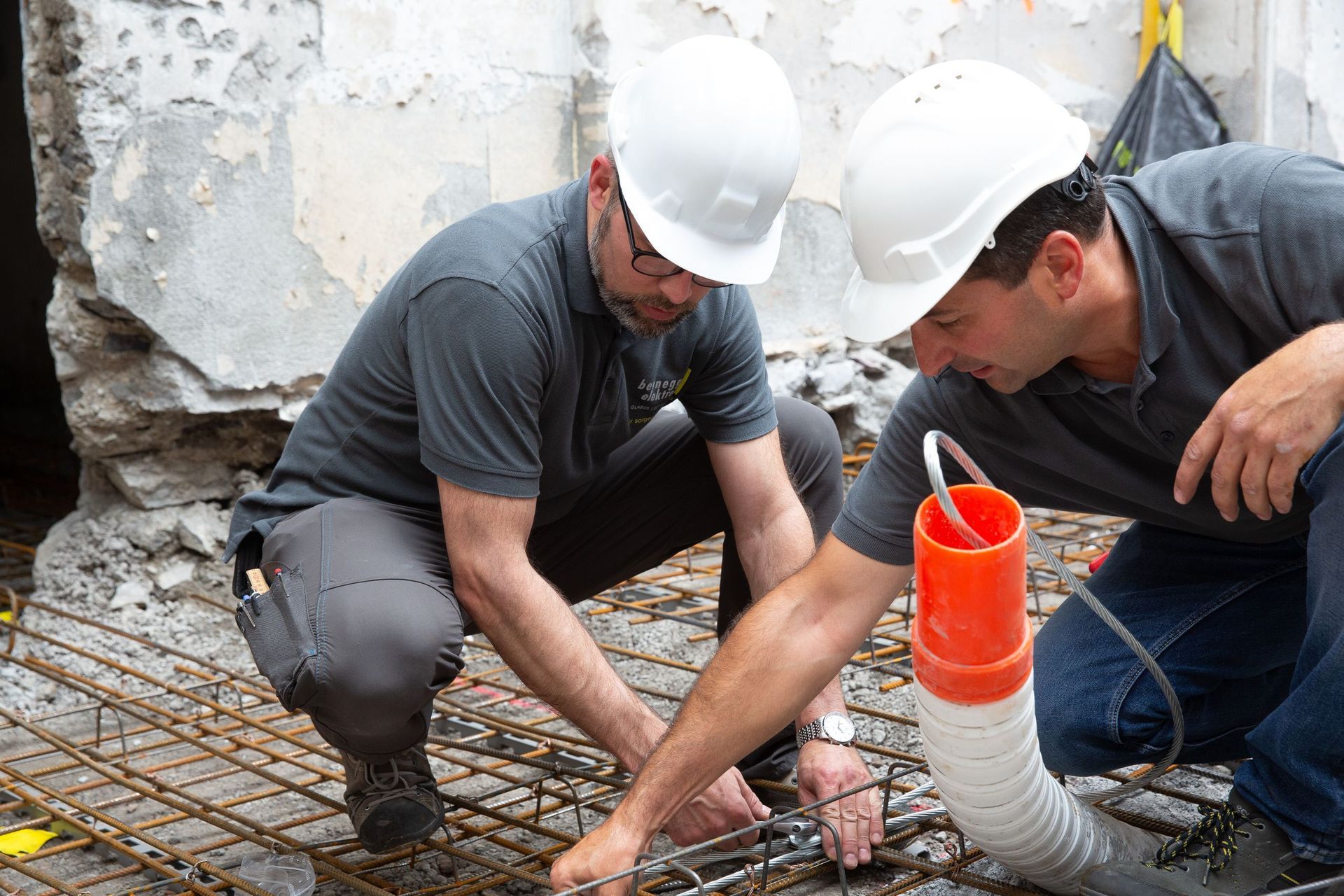 Zwei Bauarbeiter mit Schutzhelmen prüfen Bewehrungsstahl auf einer Baustelle. Einer benutzt ein Werkzeug.
