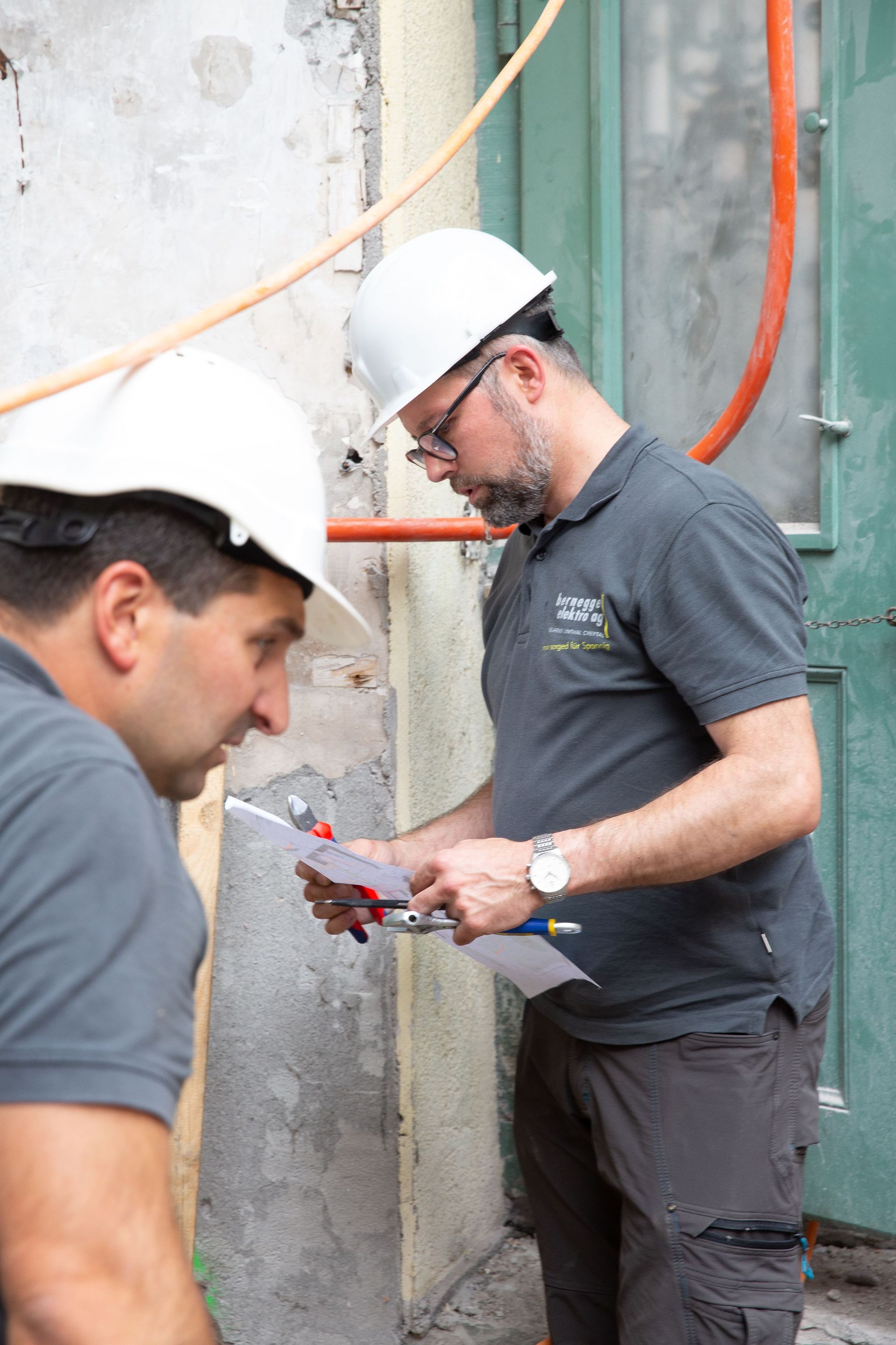 Zwei Männer mit Schutzhelmen untersuchen eine Wand. Einer hält Papiere in der Hand, der andere schaut zu. Sie stehen draußen 