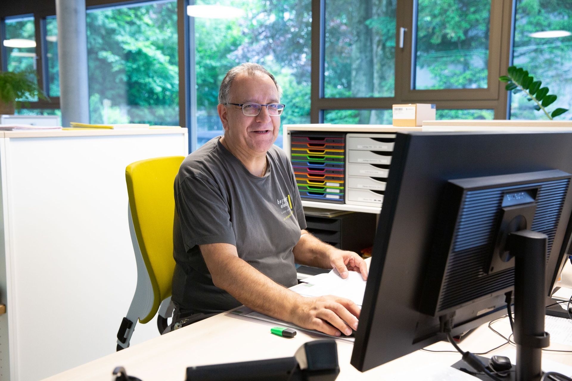 Mann mit Brille lächelt, während er in einem Büro an einem Computer arbeitet. Er trägt ein graues Hemd und sitzt an einem Sch