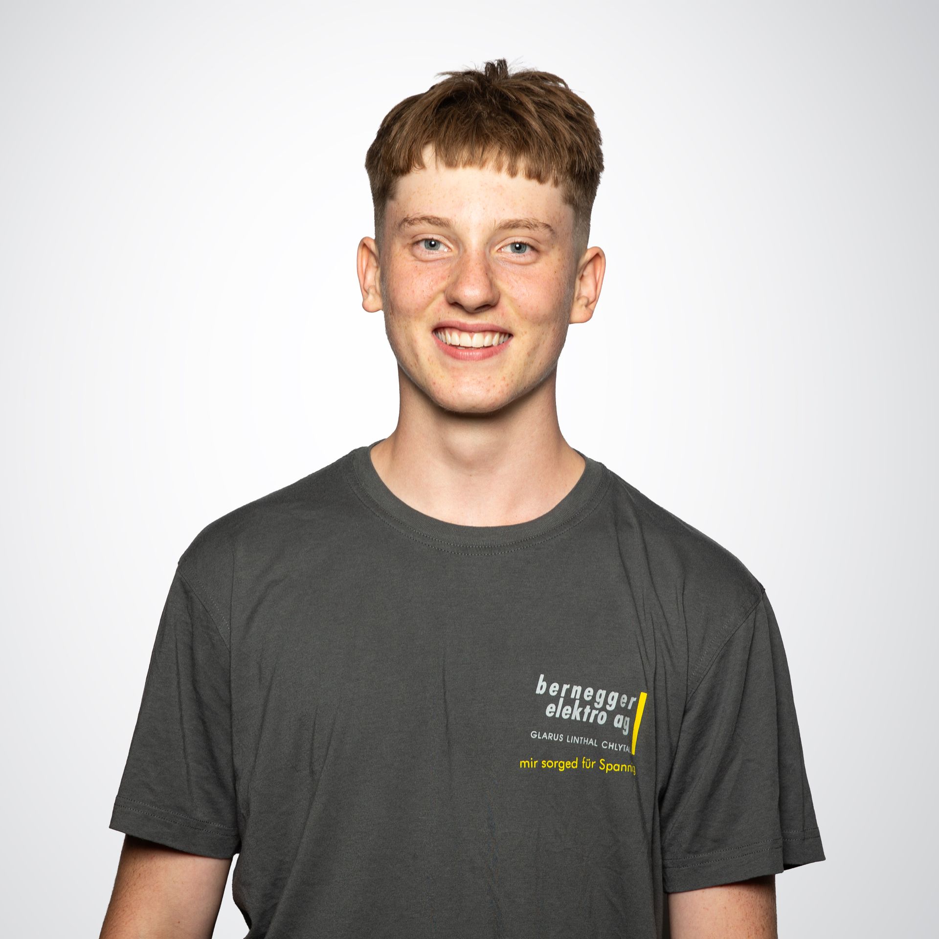 Junger Mann mit blonden Haaren und grauem T-Shirt lächelt vor weißem Hintergrund in die Kamera.
