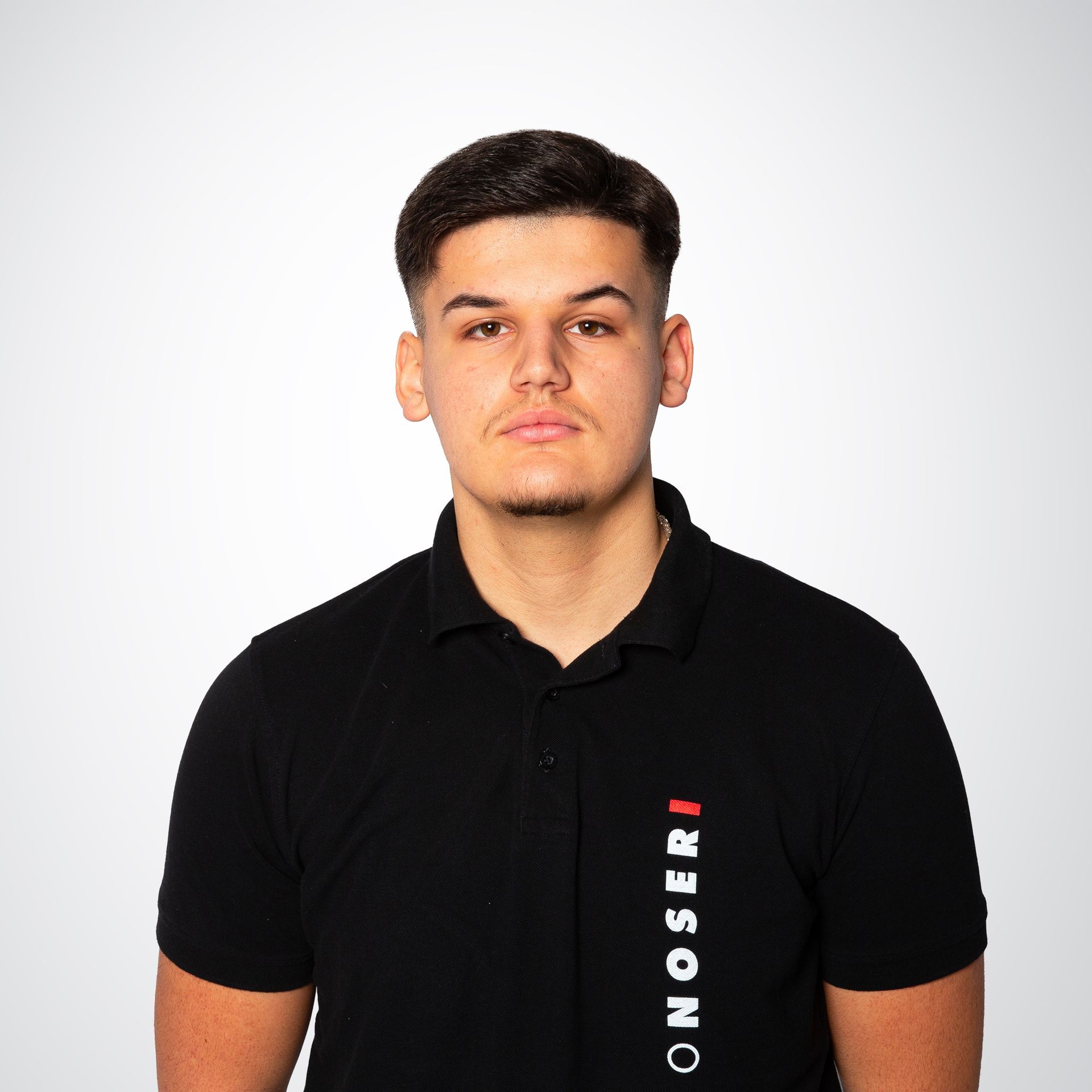 Junger Mann mit kurzen dunklen Haaren, der ein schwarzes Poloshirt mit rotem Text trägt und direkt in die Kamera blickt.