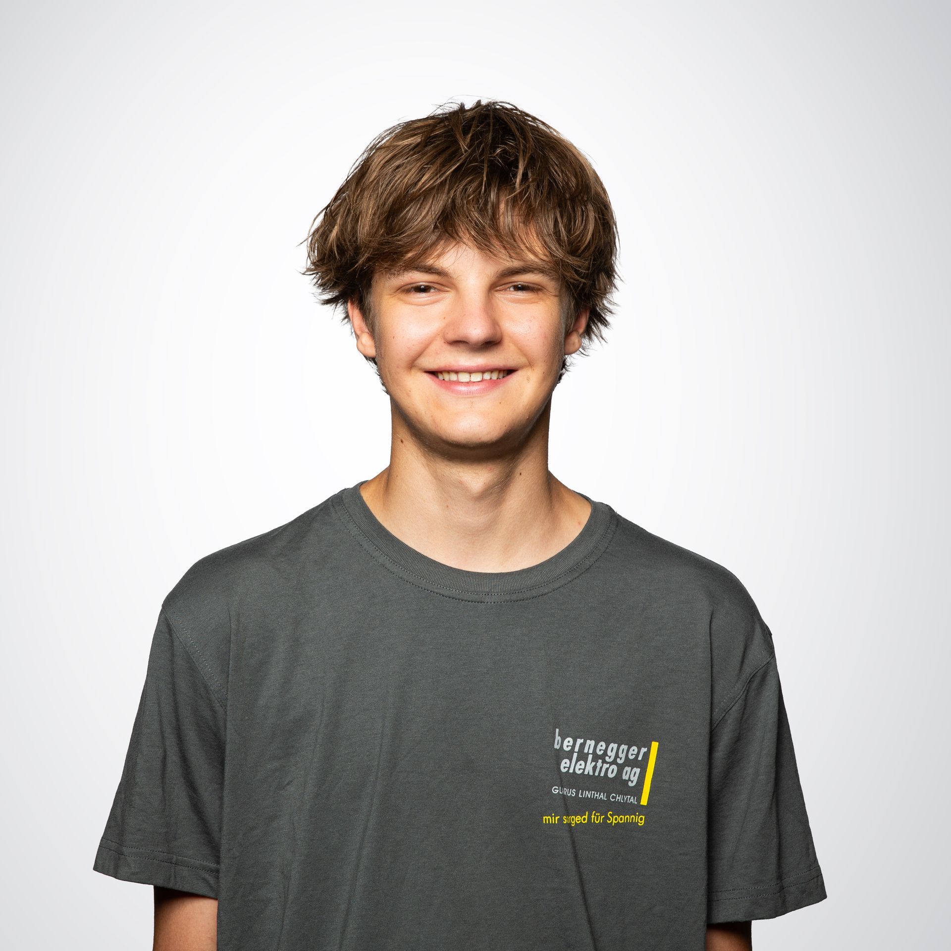Junger Mann mit blonden Haaren und grauem T-Shirt lächelt vor weißem Hintergrund in die Kamera.
