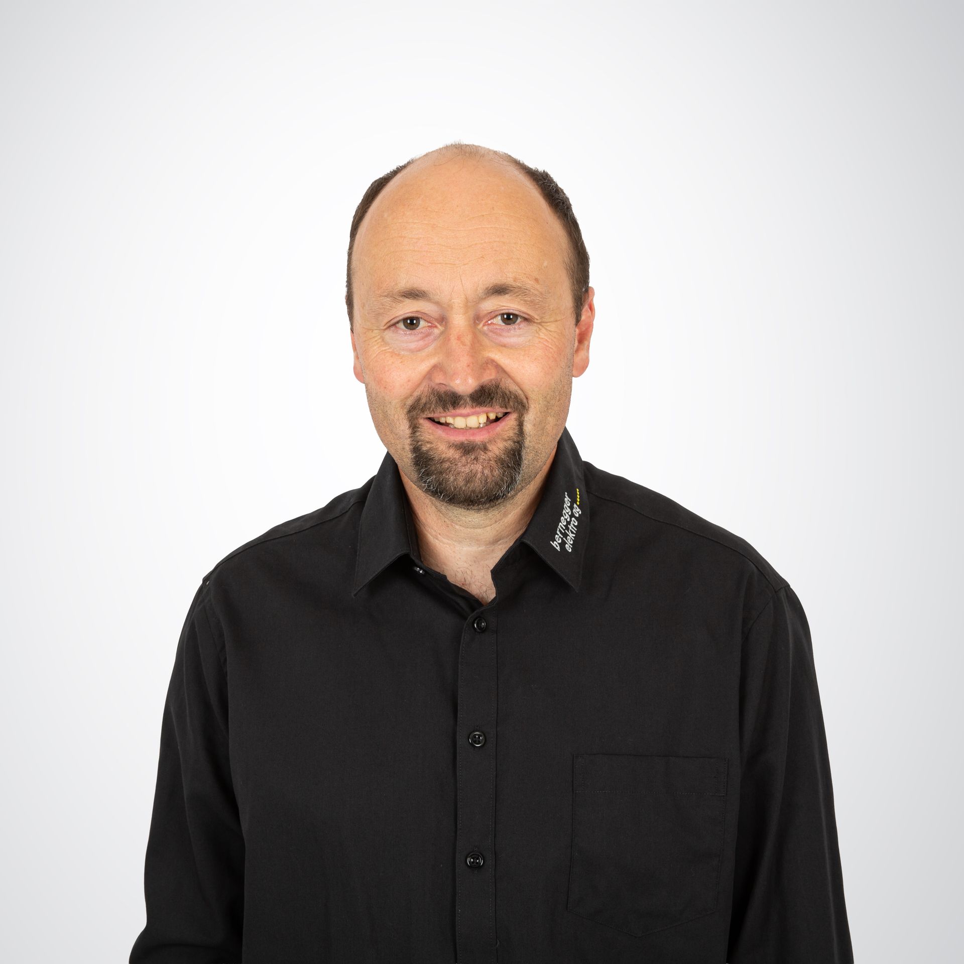 Mann mit rasiertem Kopf und Spitzbart lächelt und trägt ein schwarzes Button-Down-Hemd vor weißem Hintergrund.