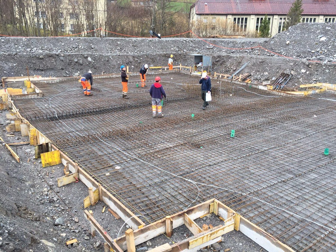 Bauarbeiter bereiten ein Fundament für den Betonguss vor. Sie stehen inmitten eines Bewehrungsgitters, umgeben von Holzschalu