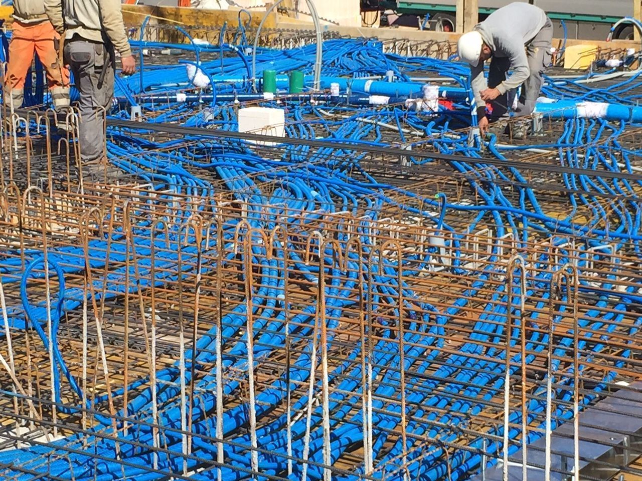 Bauarbeiter installieren blaue Rohre und Stahlbewehrung auf einem Betonfundament. Fokus auf das komplexe Materialnetzwerk.