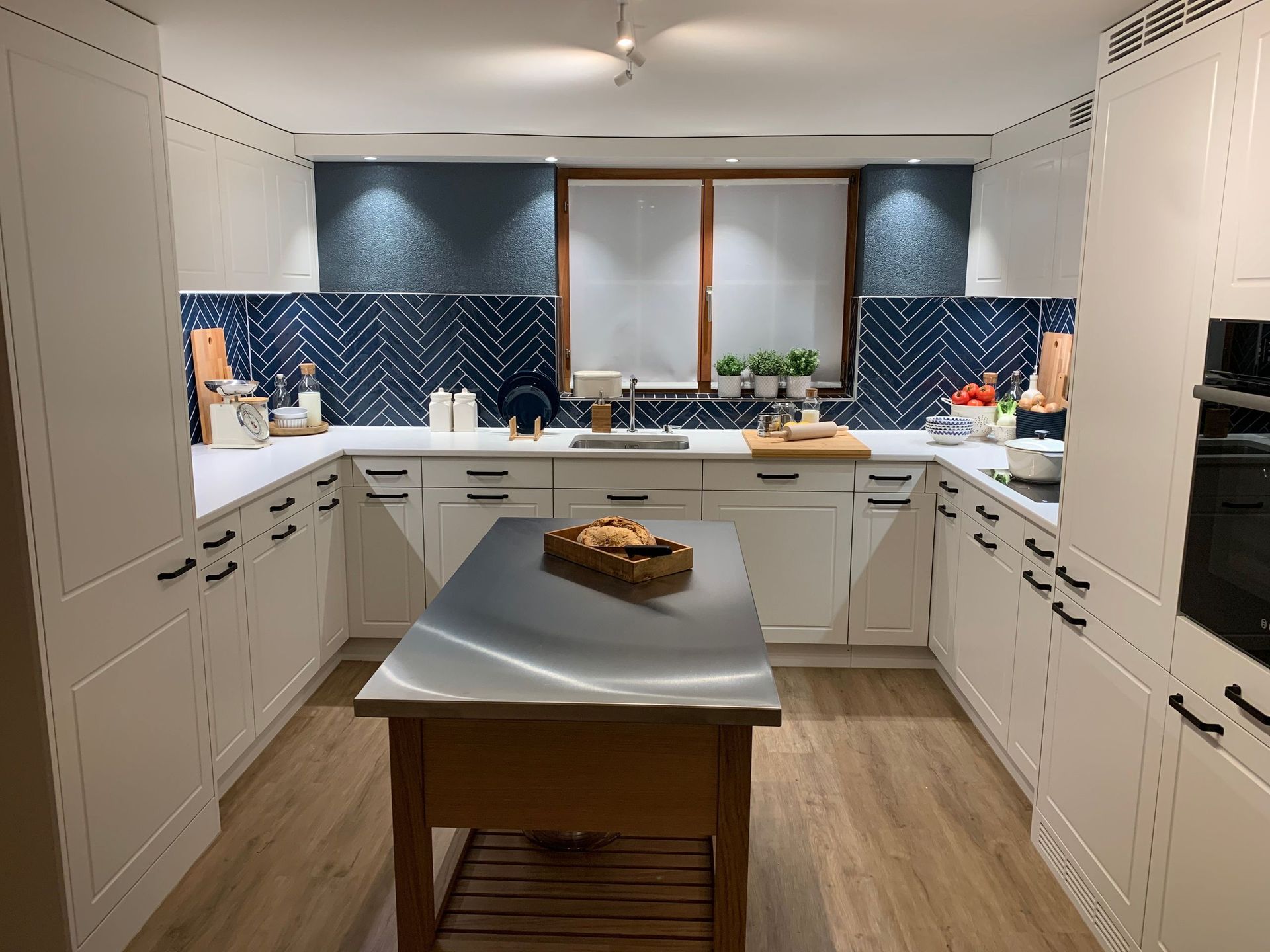 Weiße Küche mit blauer Fliesenrückwand. In der Mitte befindet sich eine Edelstahlinsel.