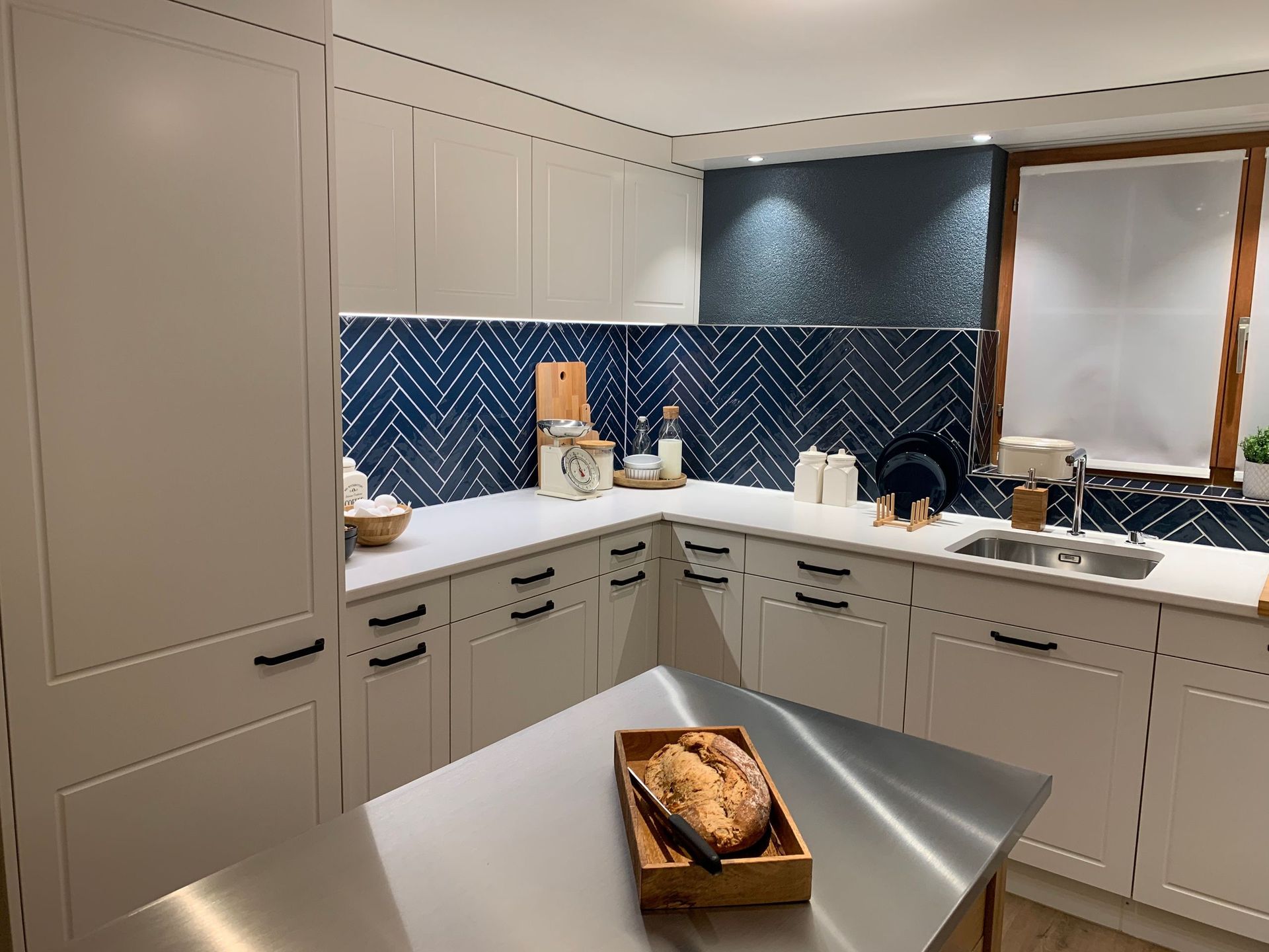 Eine moderne Küche mit weißen Schränken, blauer Fliesenrückwand und einer Edelstahlinsel. Auf der Insel liegt ein Laib Brot.