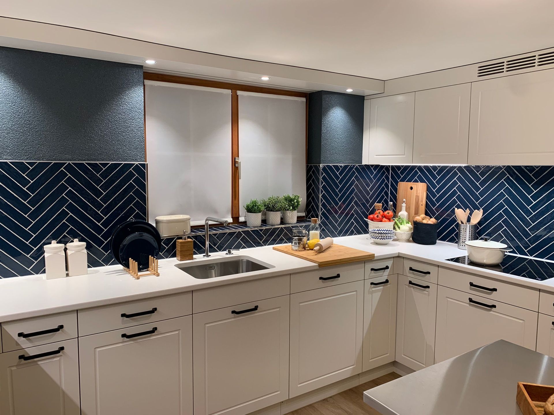 Eine moderne Küche mit weißen Schränken, blauer Fliesenrückwand und einer Edelstahlspüle. Auf der Fensterbank stehen kleine