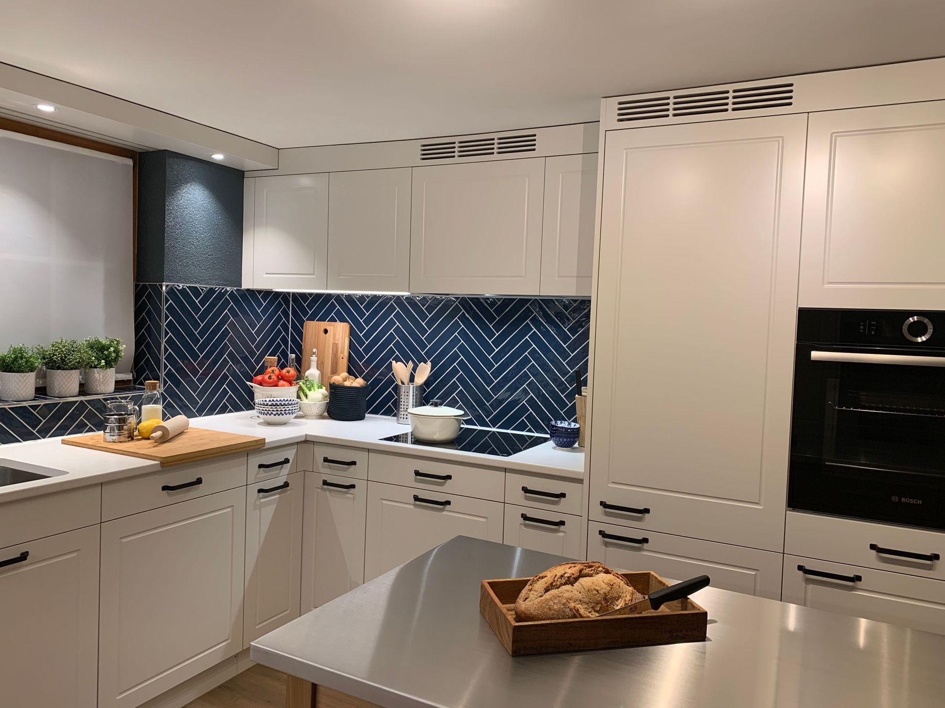 Weiße Küche mit blau gefliester Küchenrückwand. Es gibt eine Kochinsel mit Brot auf einem Tablett und einen in die