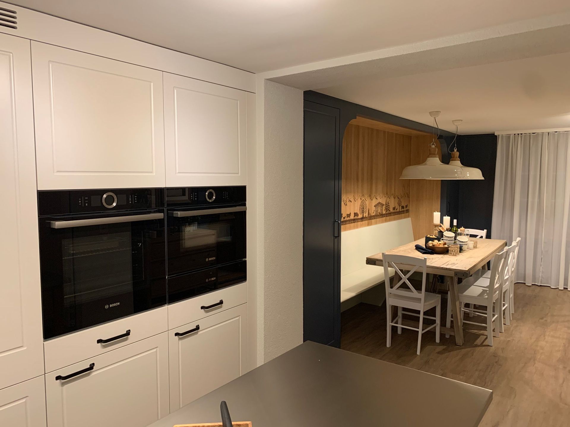 Küche mit weißen Schränken und schwarzen Geräten, die zu einem Essbereich mit einer Holzbank und einem Tisch führt.
