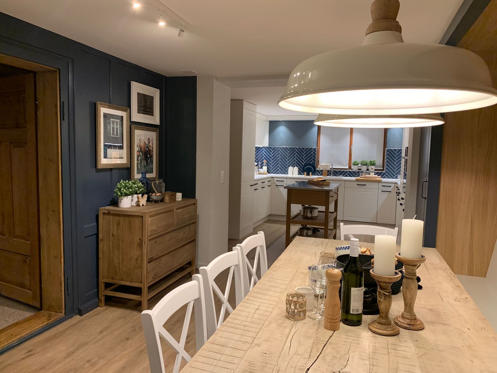 Ein Esszimmer mit einem langen, mit Kerzen gedeckten Holztisch und Blick in eine weiße Küche.