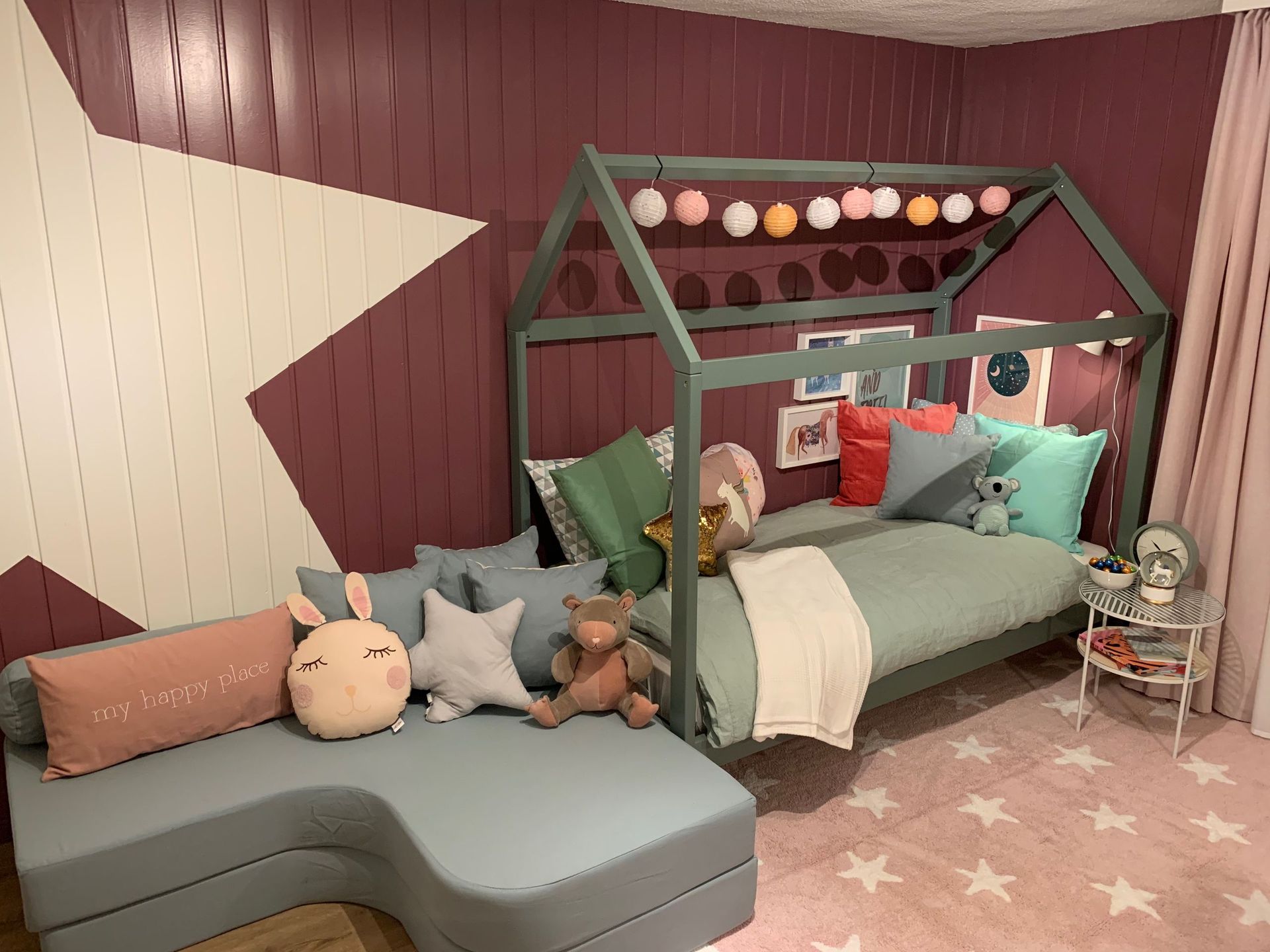 Kinderzimmer mit sternförmiger Wanddekoration, einem Bett mit Hausrahmen und einem grauen Ecksofa. Rosa und grüne Akzente.
