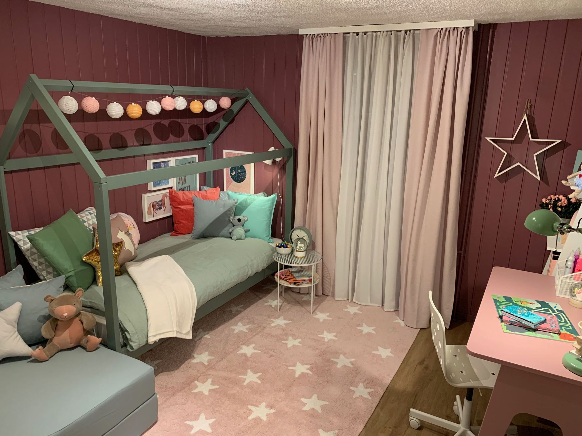 Ein Kinderzimmer in den Farben Rosa und Blaugrün. Mit hausförmigem Bettgestell, sternförmigem Teppich, Schreibtisch und Vorhä