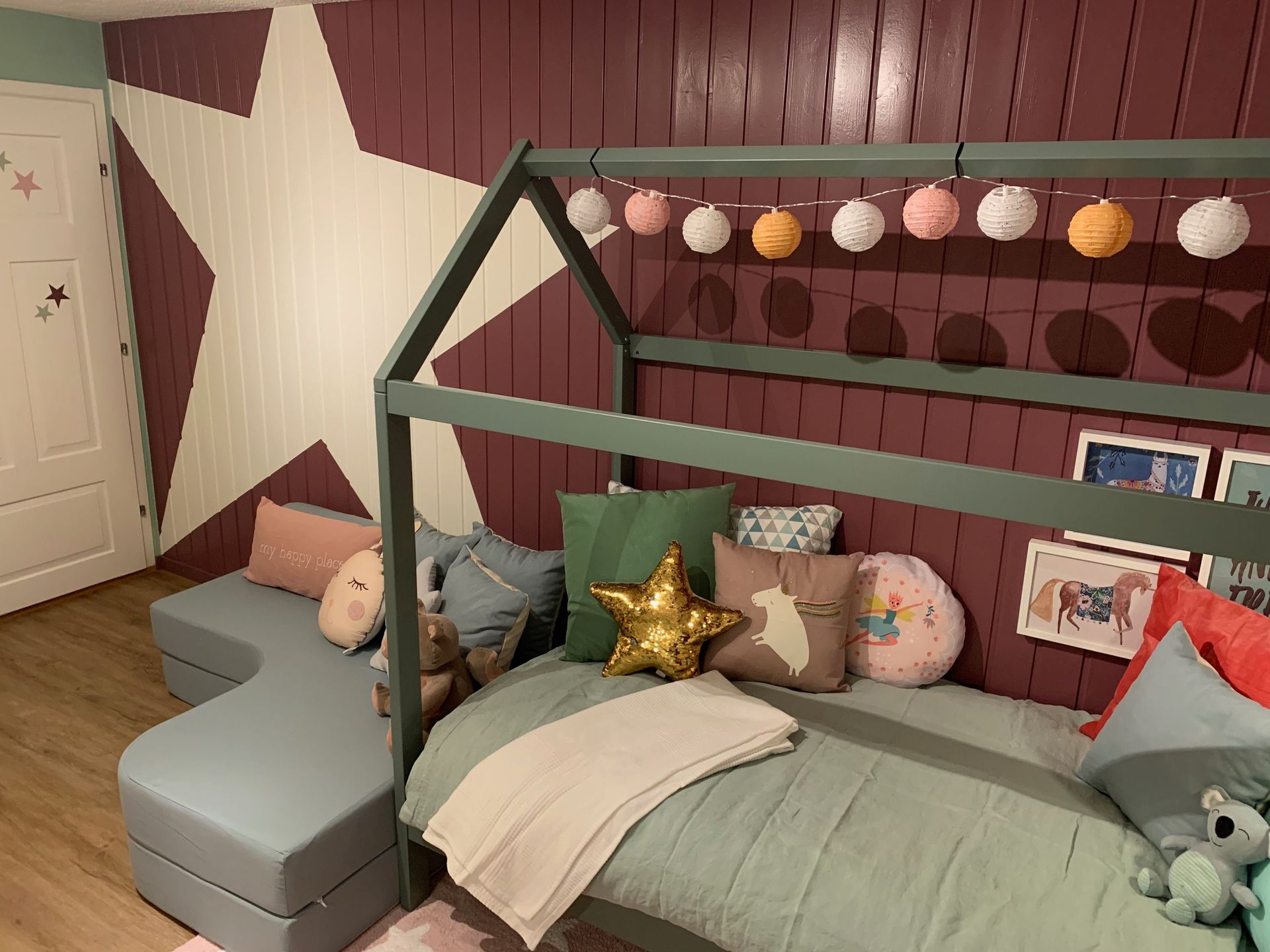 Schlafzimmer mit einem hausförmigen Bettgestell, einer mit Sternen bemalten Wand und einem grauen Sofa. Bunte Kissen und Lich