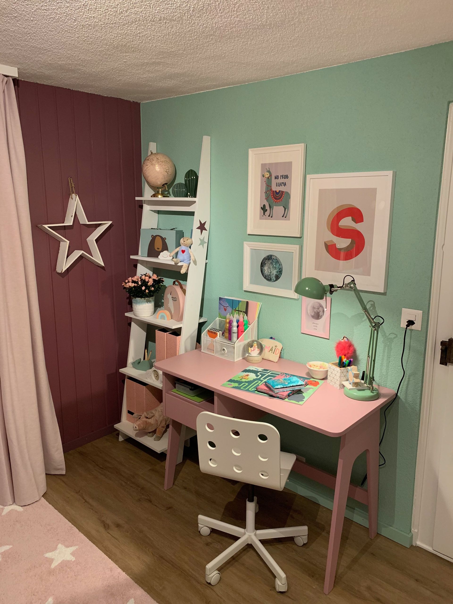 Ein pastellfarbener Kinderschreibtisch und ein Regal mit Kunstwerken und einem Stern hängen in der Ecke eines Zimmers.