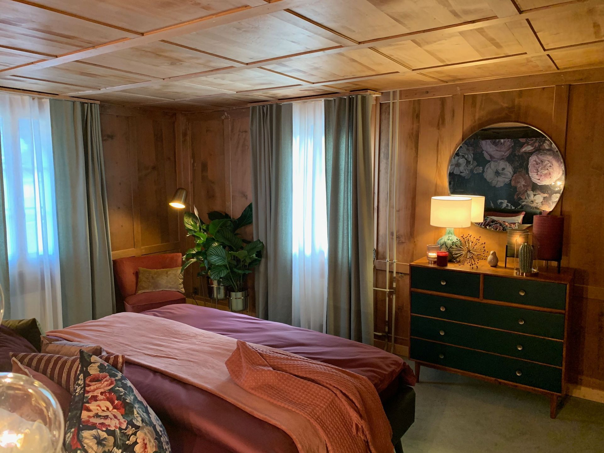 Schlafzimmer mit Holzvertäfelung, einem Bett mit rosa und lila Bettwäsche, einer grünen Kommode und einem runden Spiegel