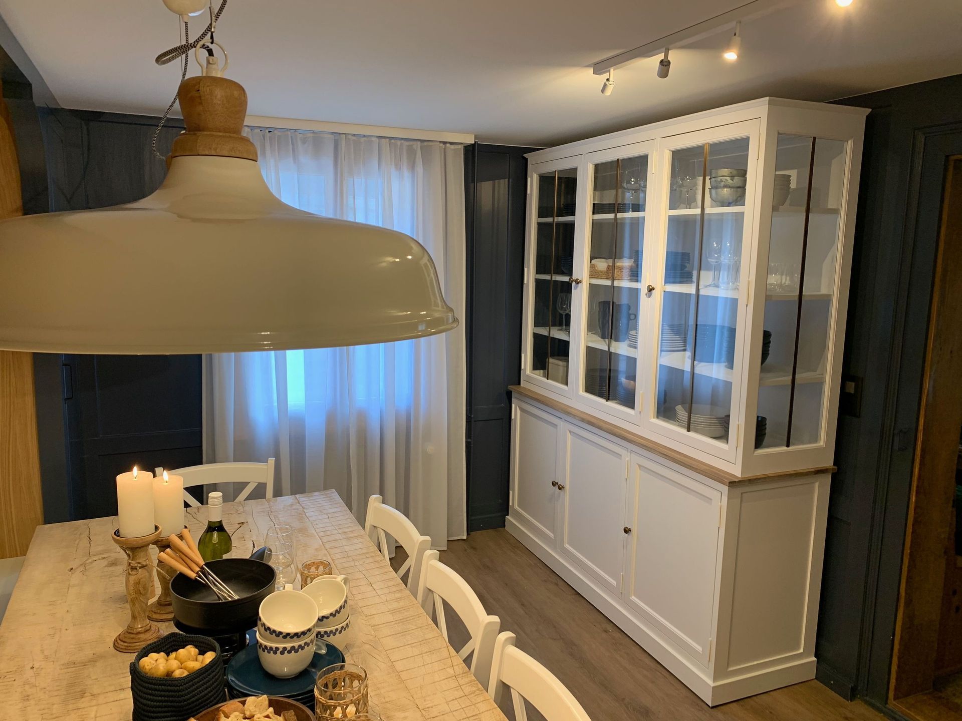 Esszimmer mit einem weißen, für eine Mahlzeit gedeckten Tisch, einer großen Hängelampe und einem weißen Schrank mit Glastüren