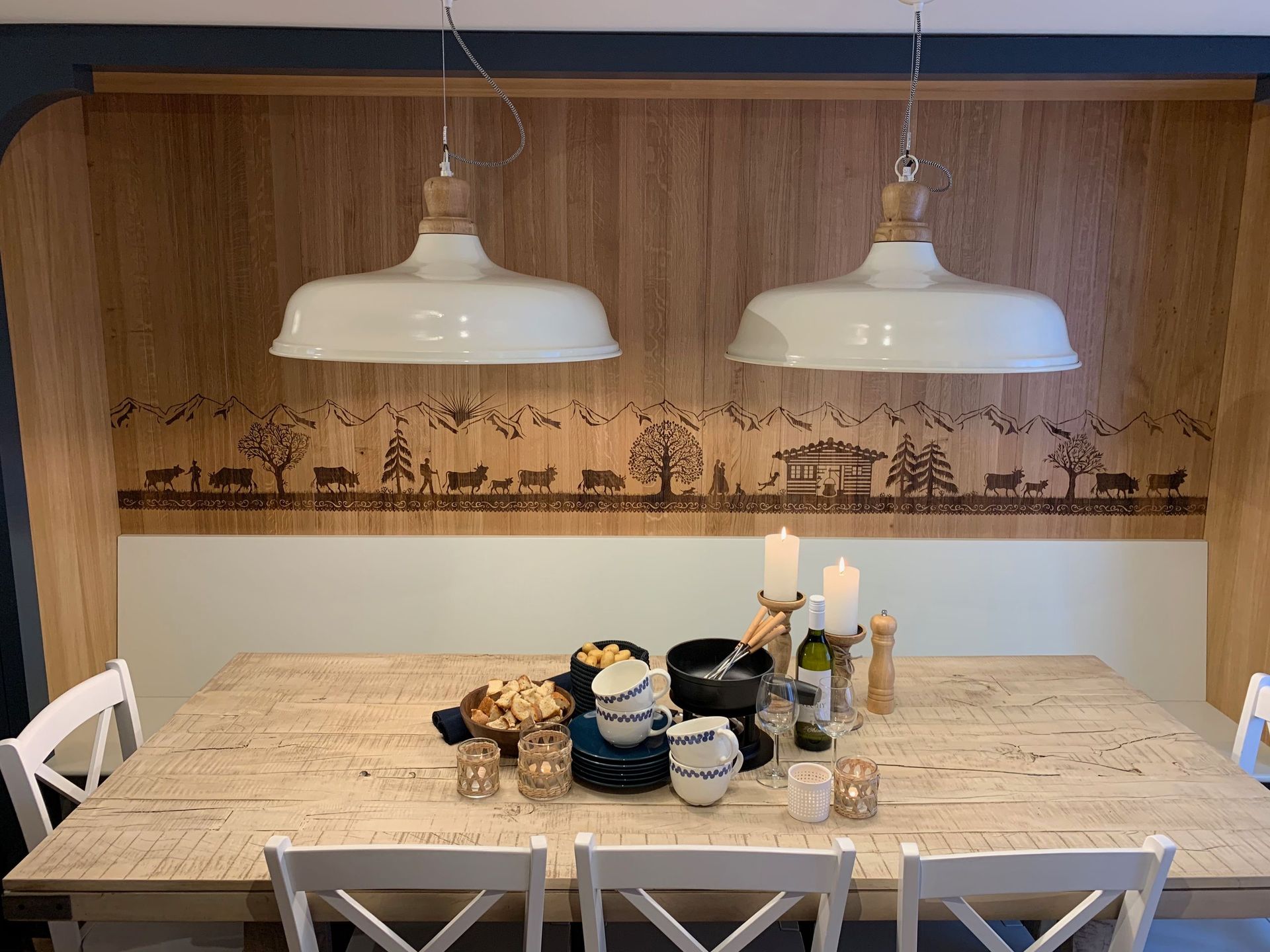 Esstisch mit weißen Stühlen, Pendelleuchten und Holzhintergrund mit einer geschnitzten Waldszene. Der Tisch ist mit