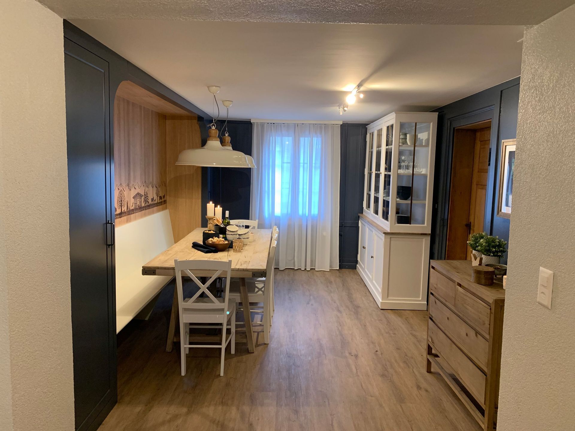 Gemütlicher Essbereich mit Holztisch, weißen Stühlen und eingebauter Bank. Dunkelblaue Wandakzente, weißer Schrank und natürl