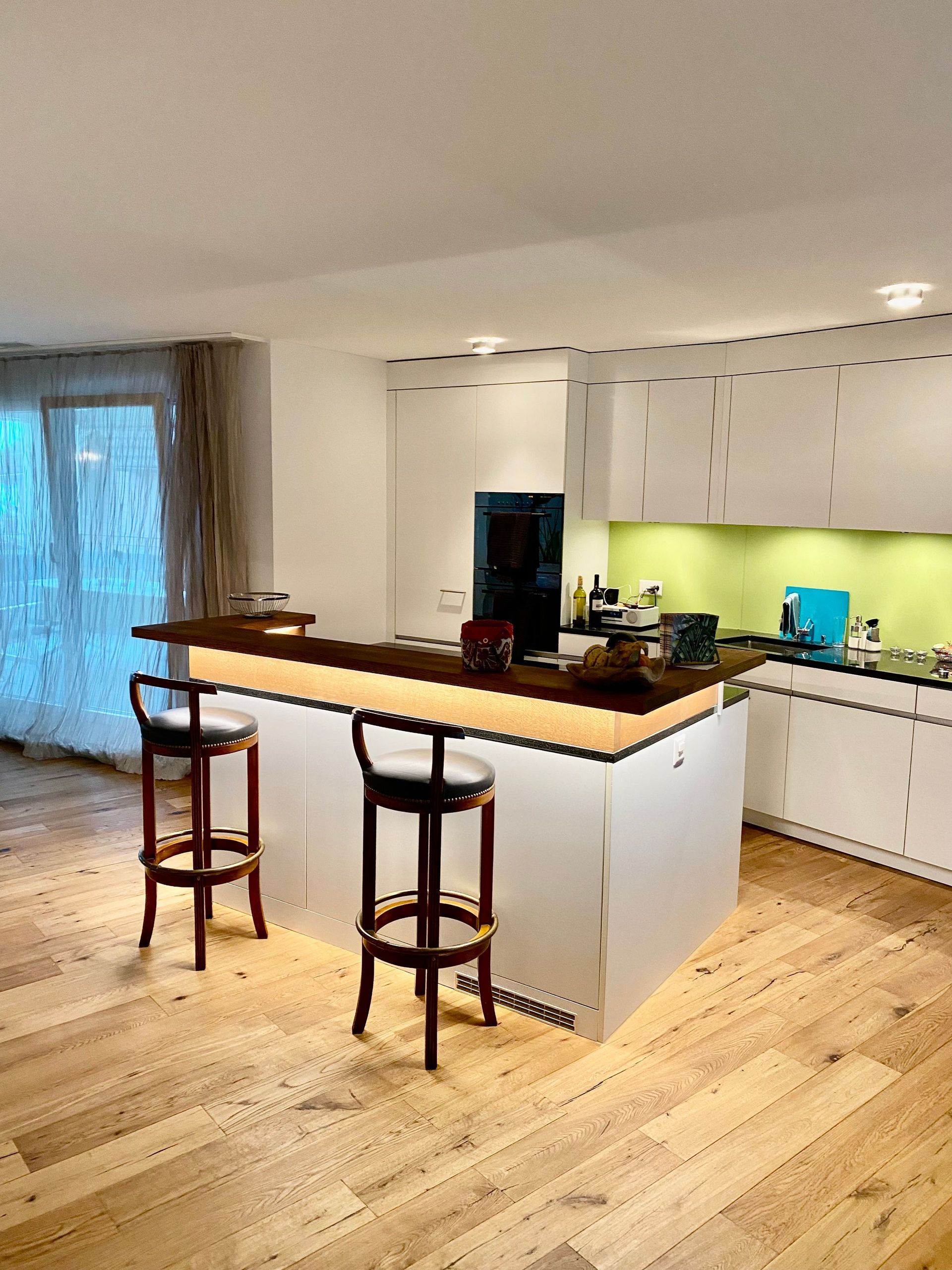 Eine moderne Küche mit einer weißen Kochinsel und Barhockern. Der Raum verfügt über Holzböden, weiße Schränke und eine dunkle