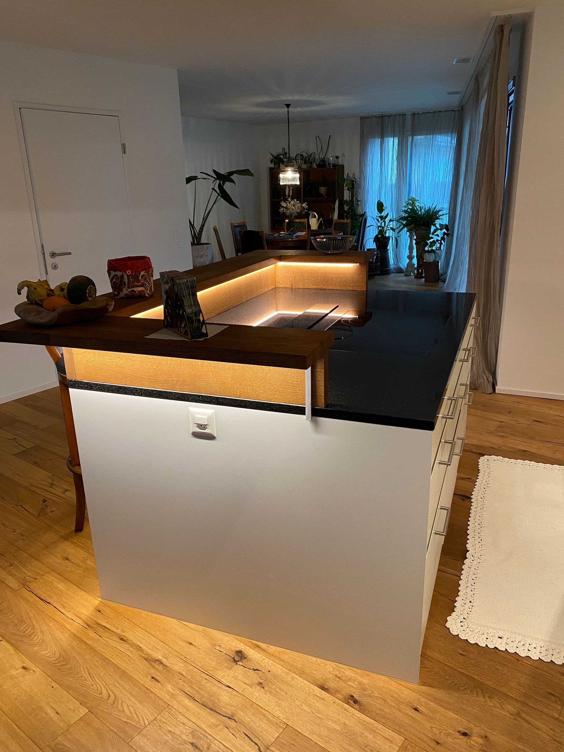 Moderne Kücheninsel mit schwarzer Arbeitsplatte, weißen Schränken und warmen Holzakzenten. Integrierte Beleuchtung erhellt di