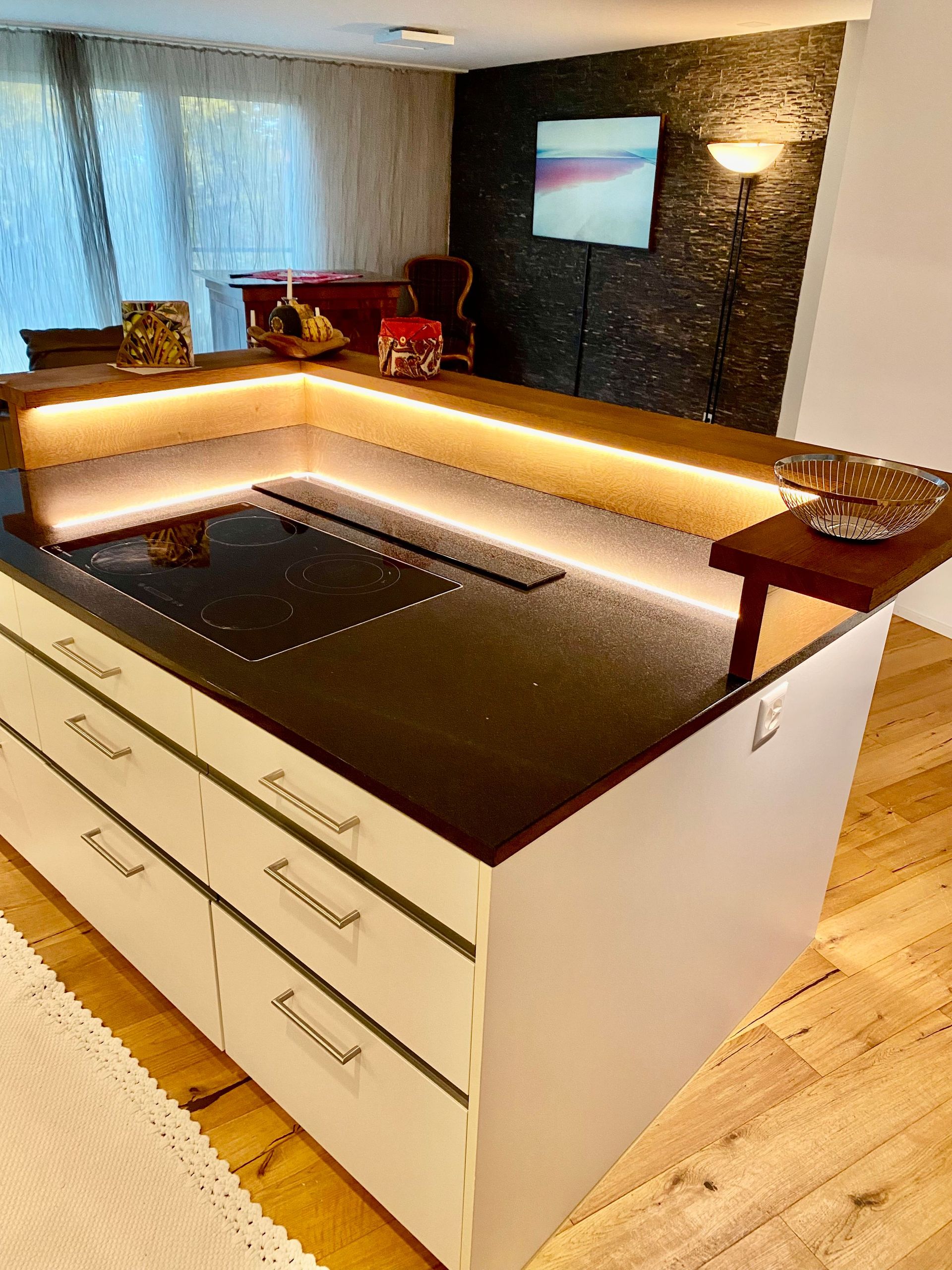 Moderne Kücheninsel mit weißen Schränken, dunkler Arbeitsplatte mit eingebautem Kochfeld und Streifenbeleuchtung, im Hintergr