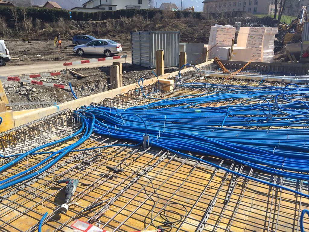 Baustelle mit blauen Rohren, die über einem Metallgitter verlegt und zum Betonieren vorbereitet sind. Im Hintergrund sind Aut