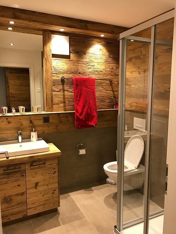 Rustikales Badezimmer mit Holzvertäfelung, rotem Handtuch, Glasdusche und weißer Toilette.