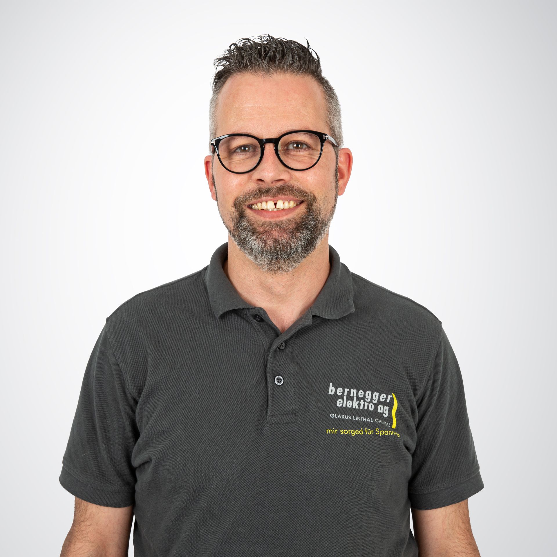 Mann mit Brille und ergrauendem Haar, trägt ein graues Poloshirt und lächelt vor einem weißen Hintergrund.