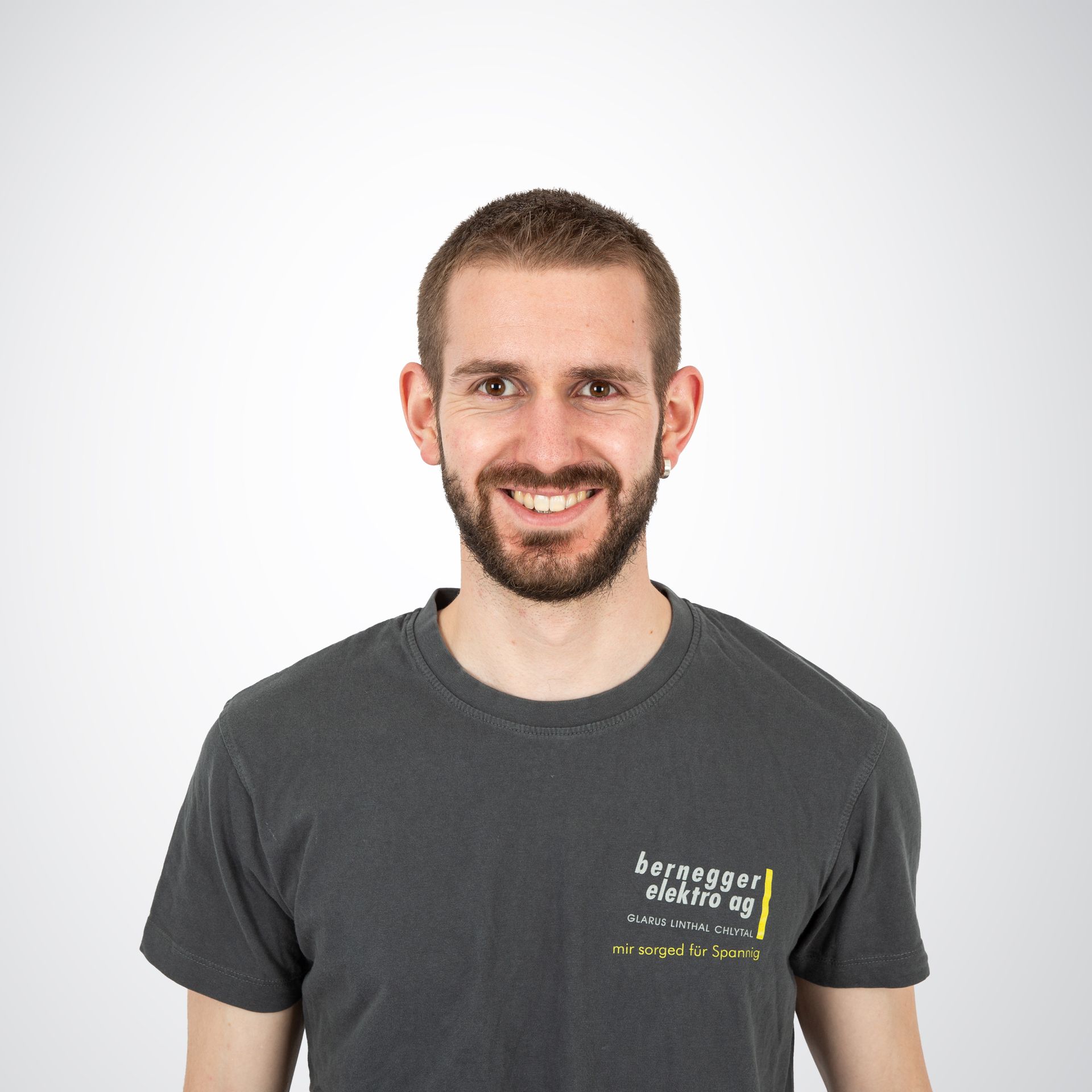Mann in grauem T-Shirt mit Logo, lächelt in die Kamera.