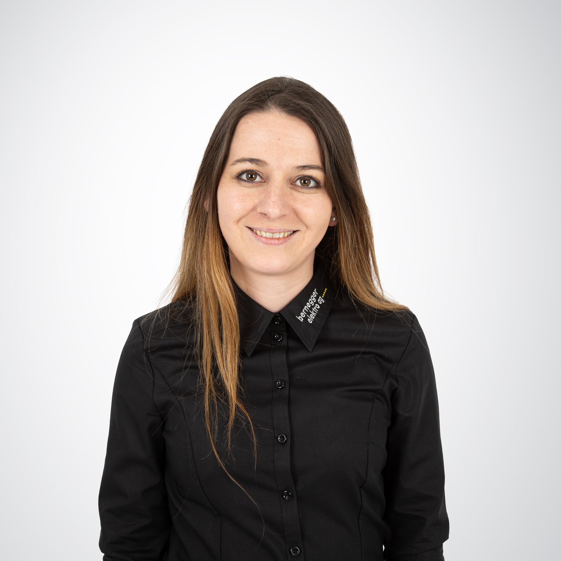 Frau mit langen braunen Haaren, trägt ein schwarzes Button-Down-Hemd und lächelt vor einem weißen Hintergrund.