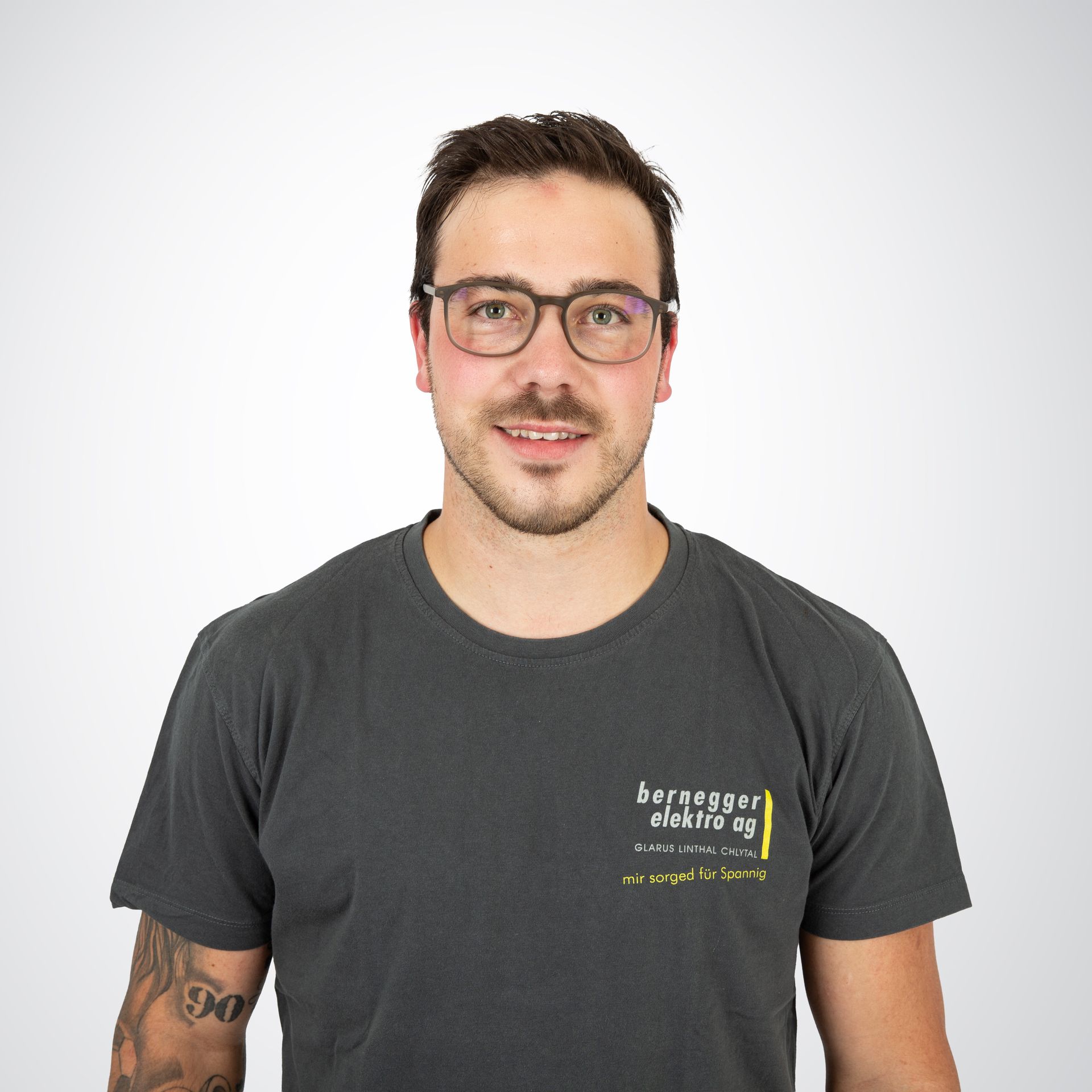 Mann mit Brille und grauem T-Shirt mit Logo, lächelnd, die Arme an den Seiten, vor weißem Hintergrund.
