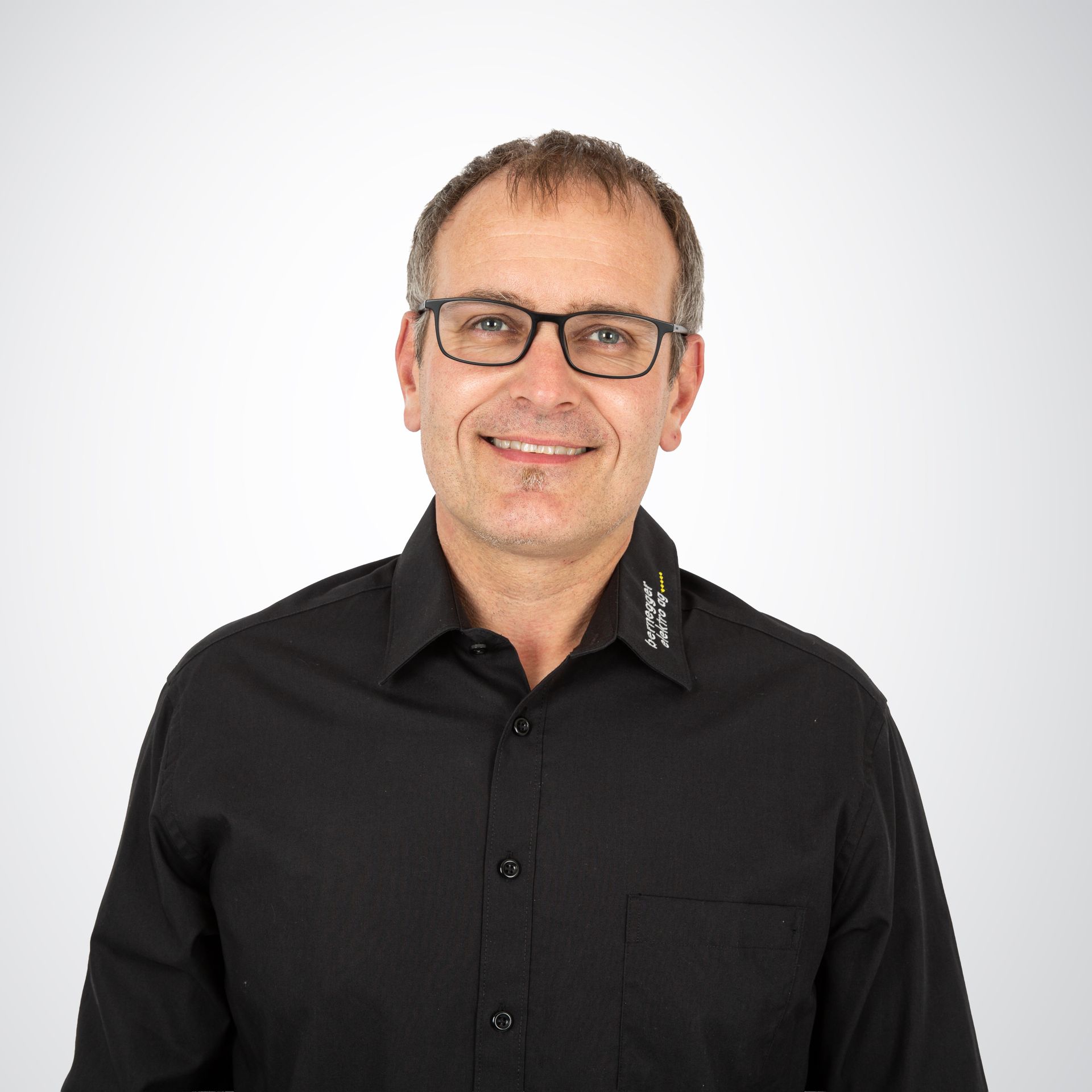 Lächelnder Mann mit Brille, in schwarzem Button-Down-Hemd vor weißem Hintergrund.