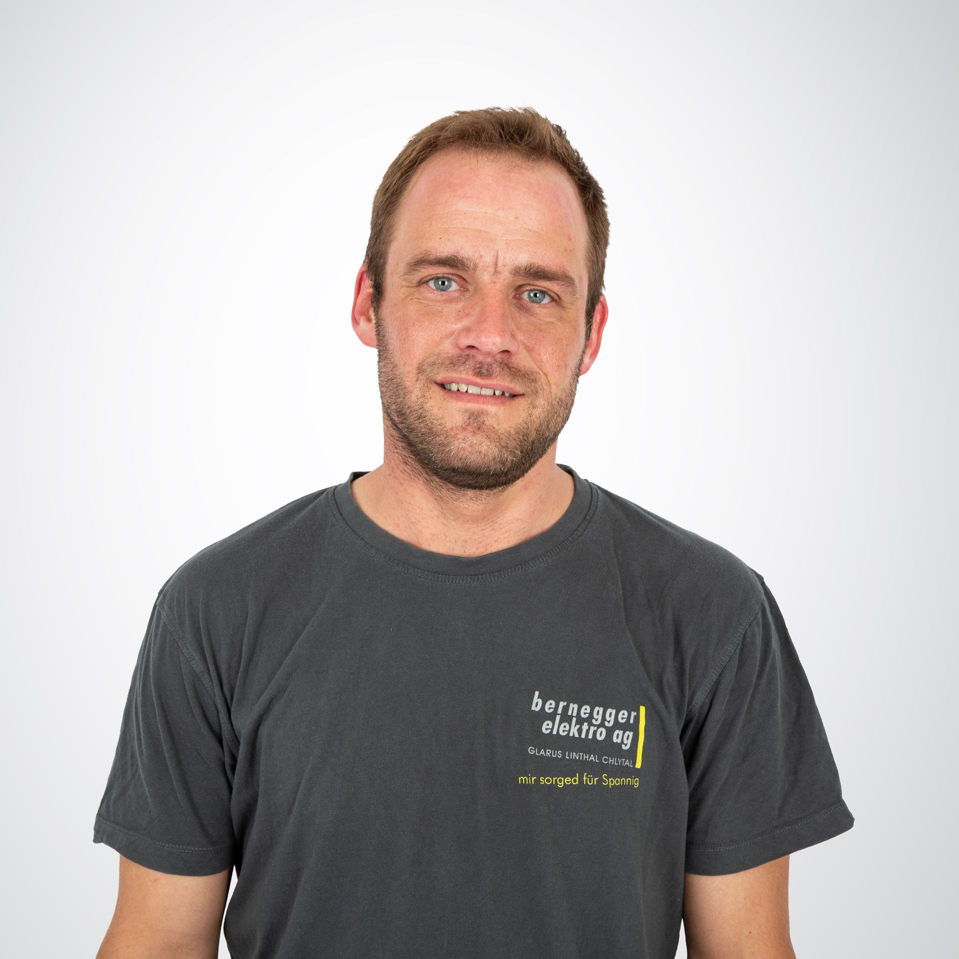 Ein Mann in einem grauen T-Shirt mit einem Logo lächelt in die Kamera und steht vor einem weißen Hintergrund.