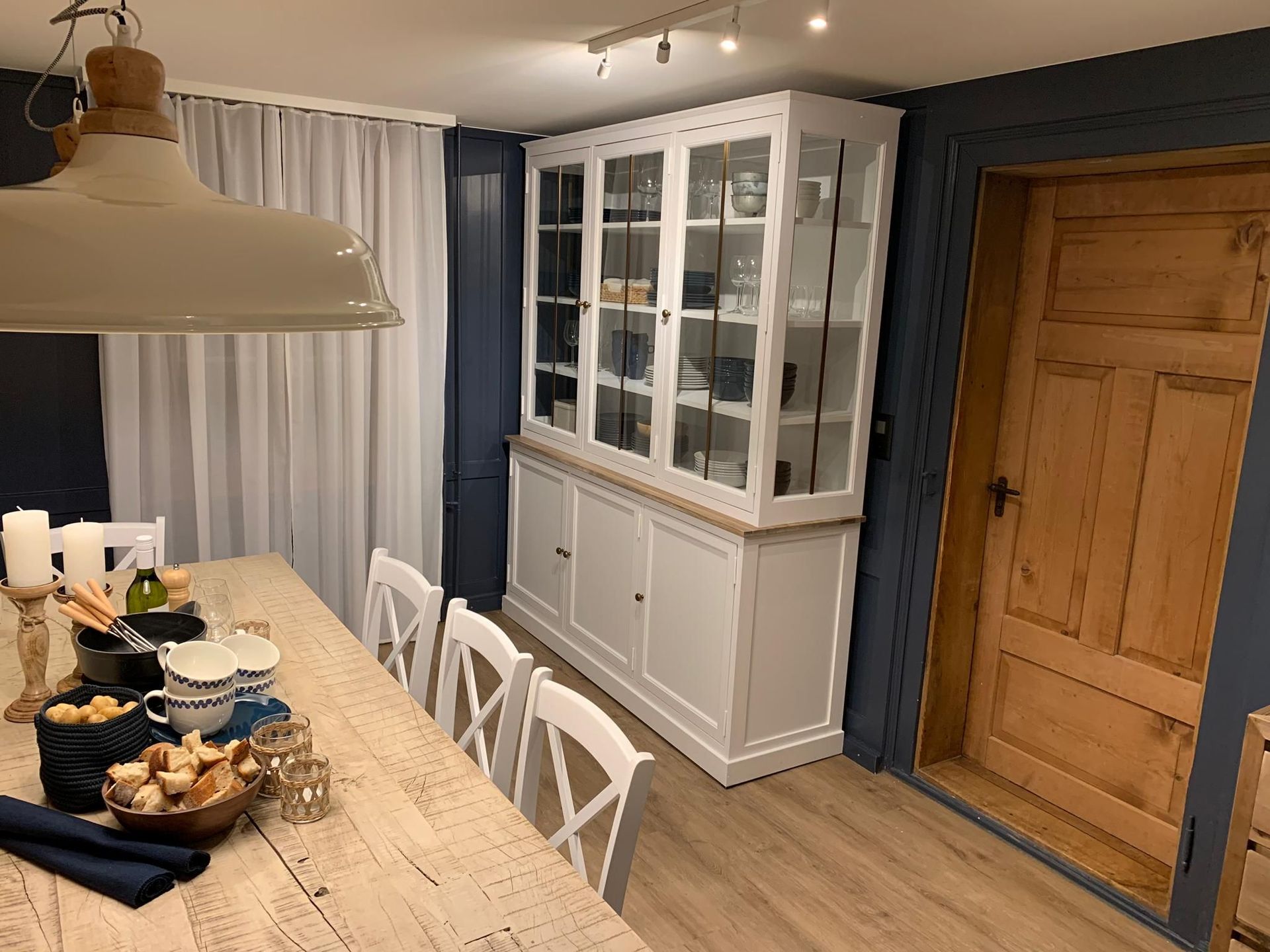 Esszimmer mit weißem Schrank, gedecktem Holztisch und rustikaler Holztür vor marineblauen Wänden.