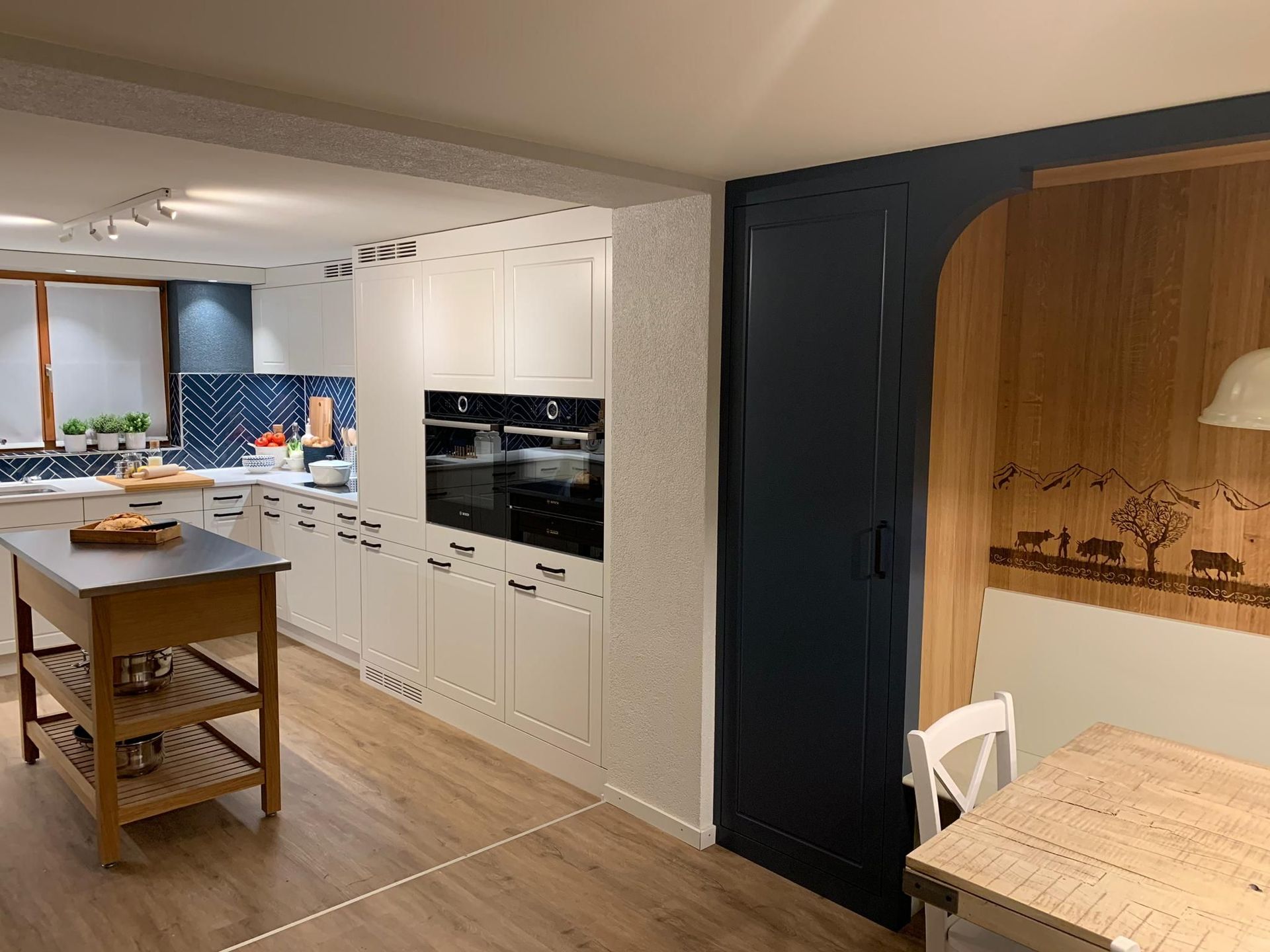 Küche mit weißen Schränken, Kochinsel und Backofen. Rechts befindet sich eine dunkelblaue Tür mit Holzvertäfelung, die zum Es