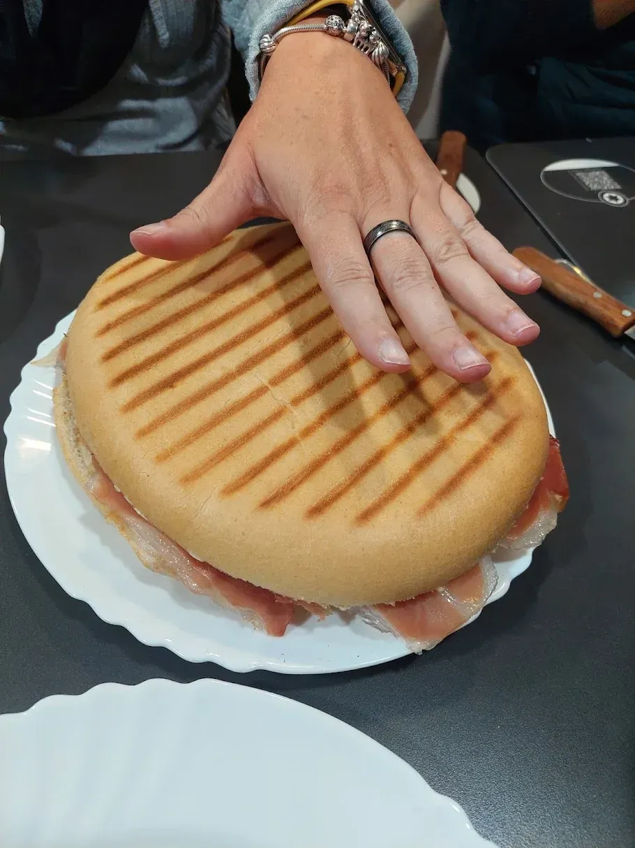 Mano presionando un sándwich grande con pan tostado, jamón y un plato blanco sobre una mesa.