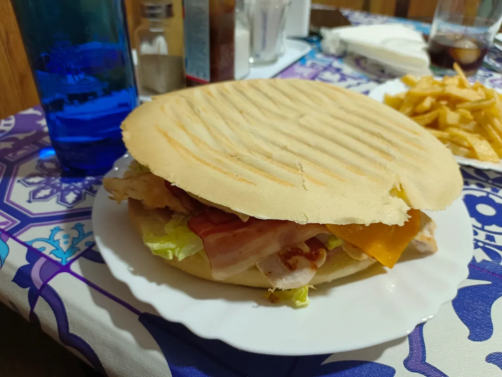 Sándwich con bacon, lechuga y queso en un plato, junto con papas fritas, una bebida y un vaso de agua