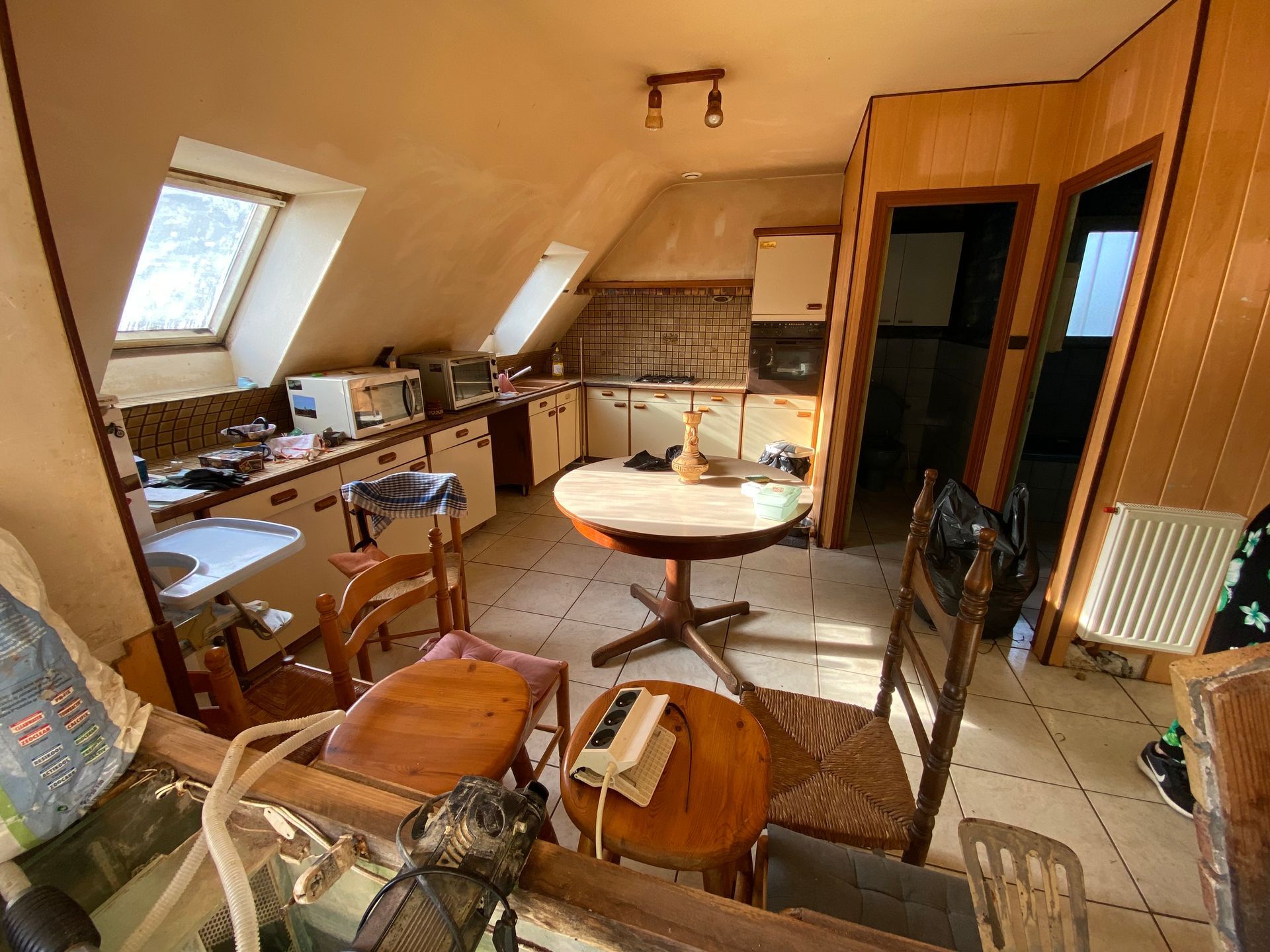 Cuisine encombrée avec plafond mansardé, appareils électroménagers, table et chaises.