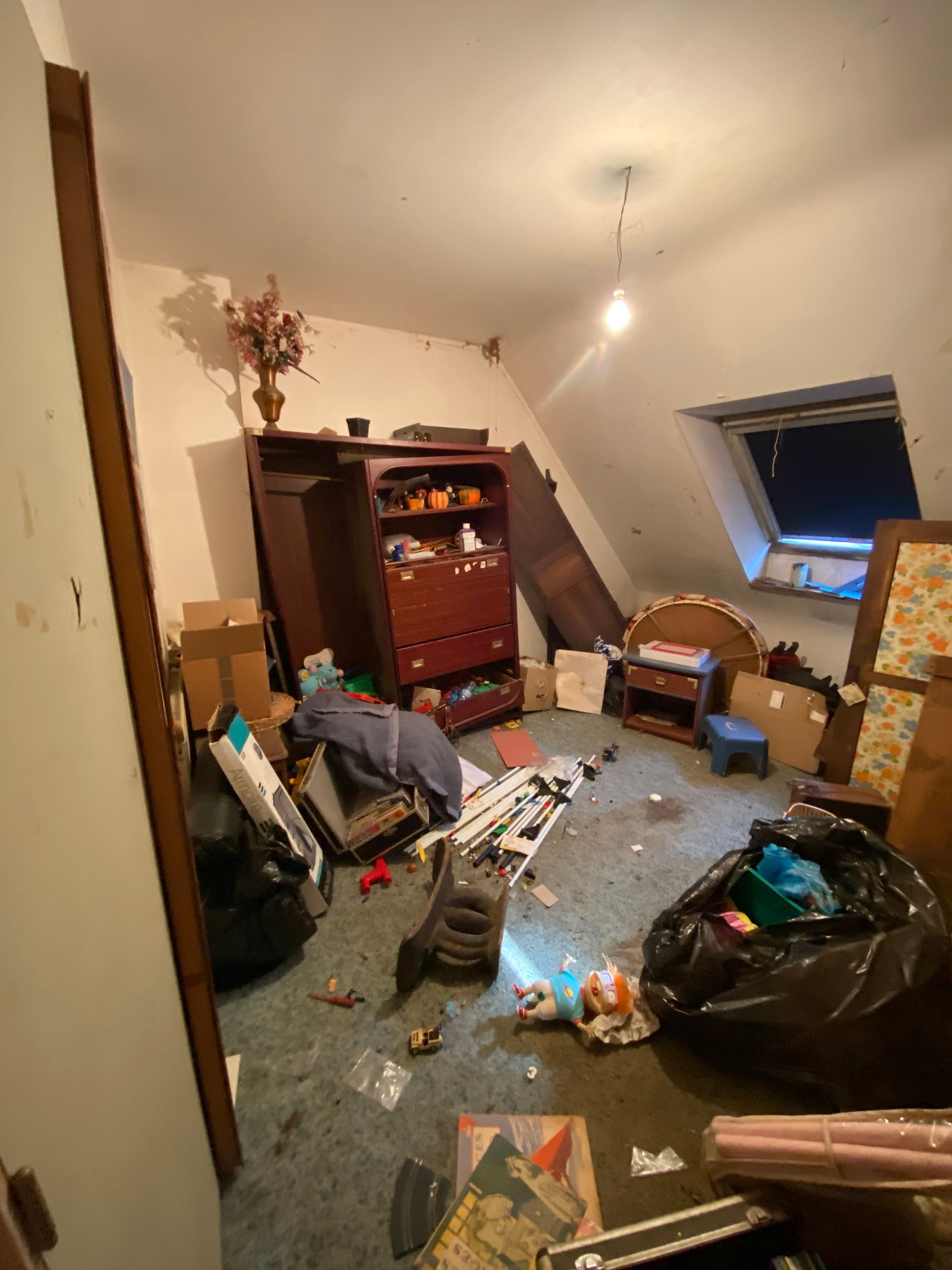 Pièce en désordre avec beaucoup de choses : meubles, sacs poubelles, débris au sol et un plafond mansardé avec une fenêtre.