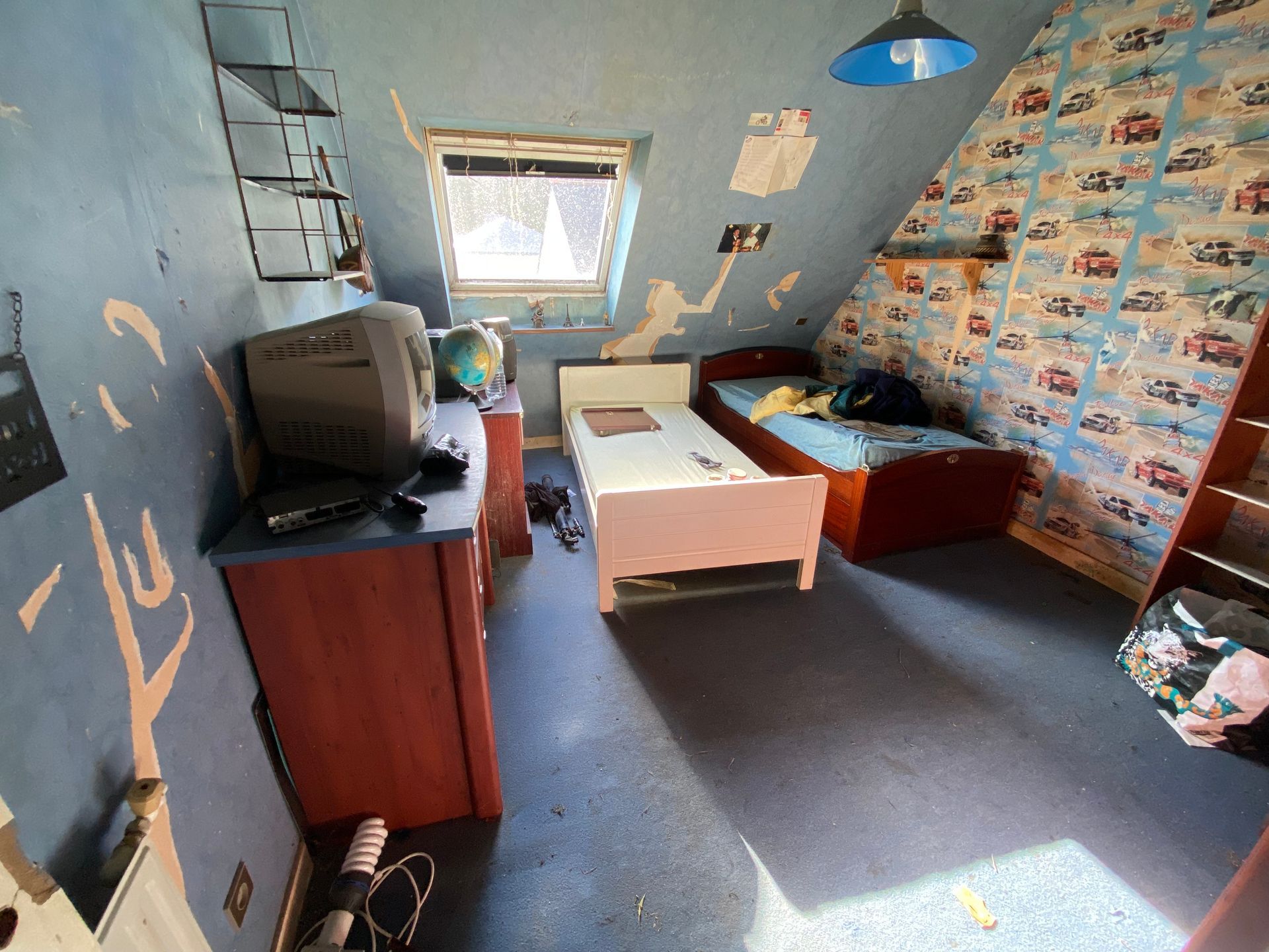 Chambre d'enfant avec papier peint bleu qui se décolle, deux lits et une télévision sur une commode.