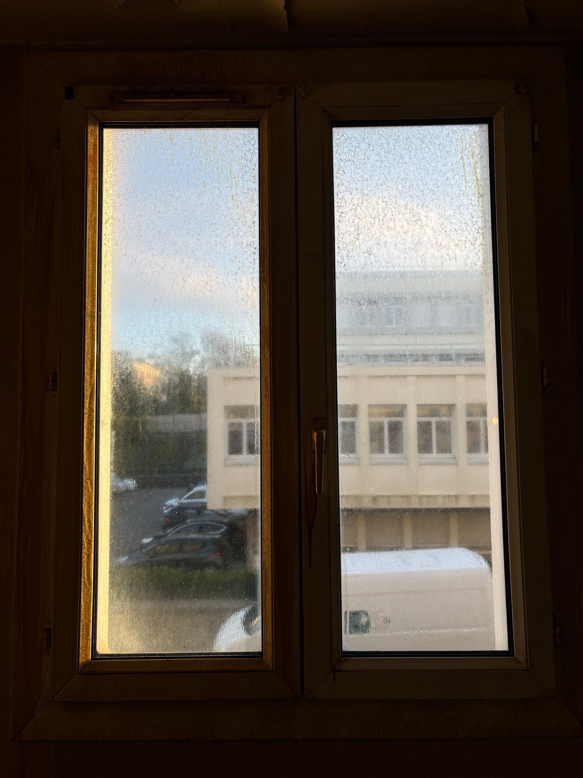 Fenêtre embuée, donnant sur un bâtiment et des voitures garées à l'extérieur par temps de pluie.