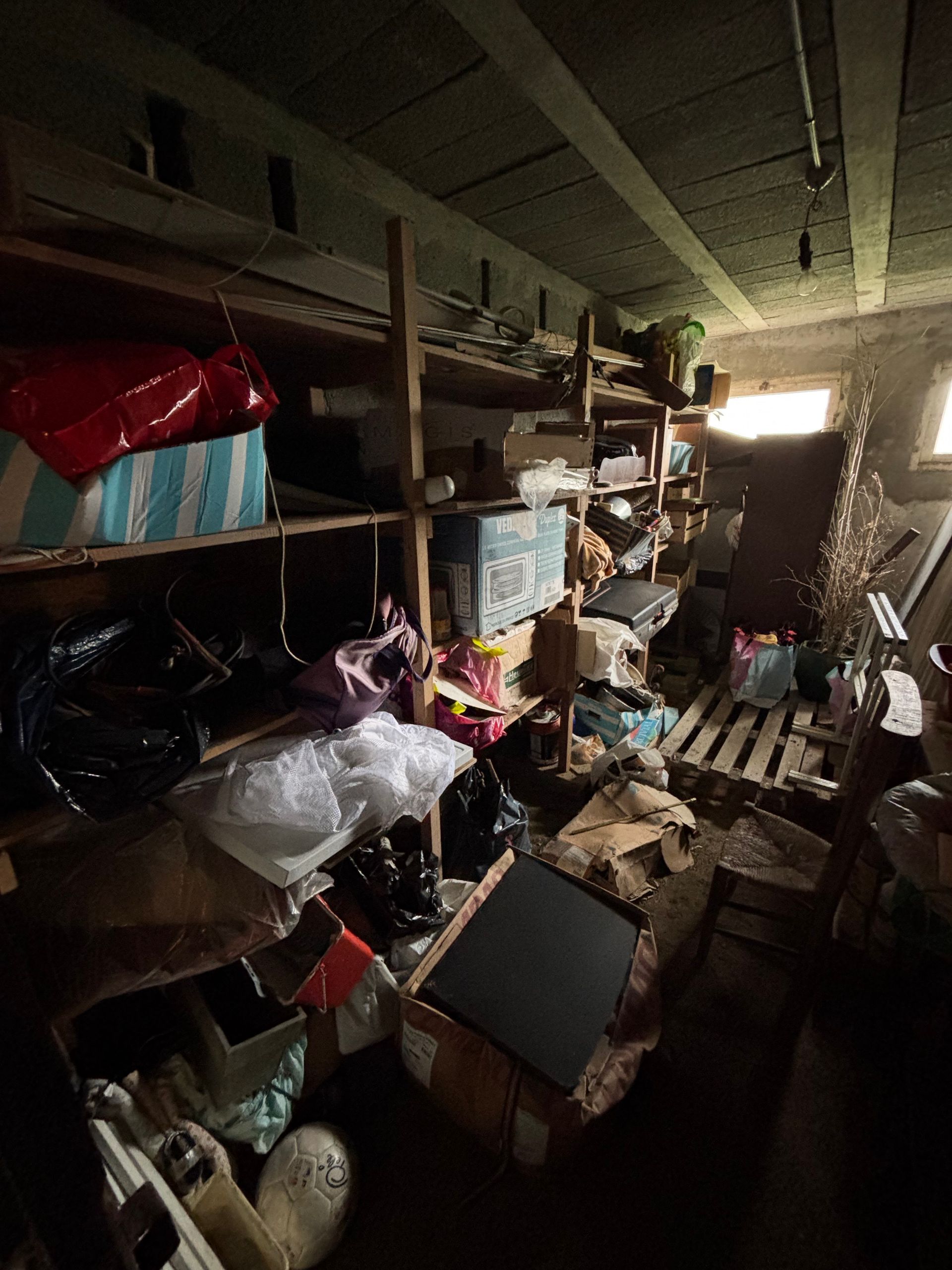 Débarras encombré, avec des étagères en bois débordant de cartons, de sacs et d'objets divers. Éclairage tamisé.