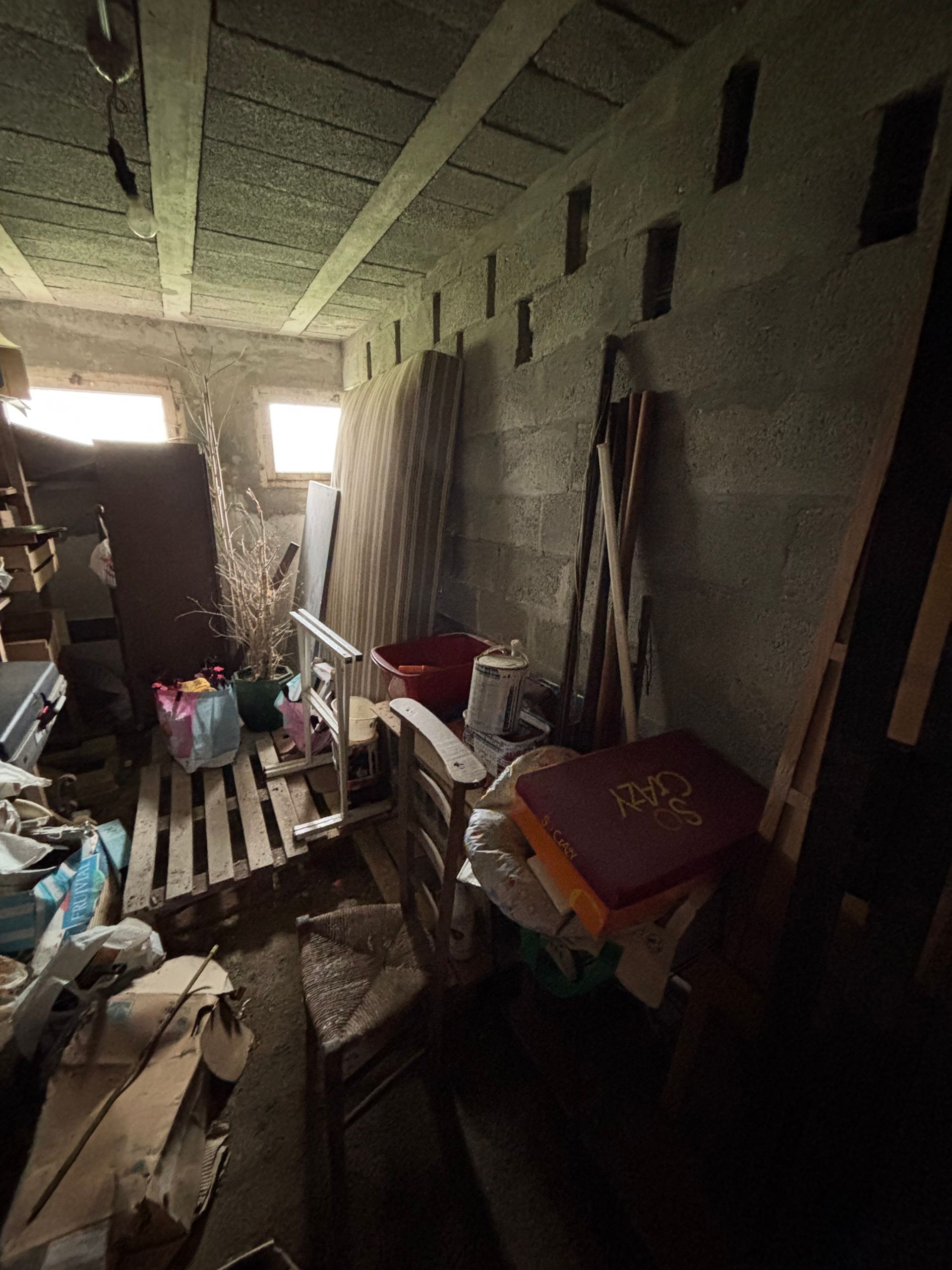 Un débarras encombré, avec des murs en béton, des poutres apparentes au plafond et divers objets entreposés.